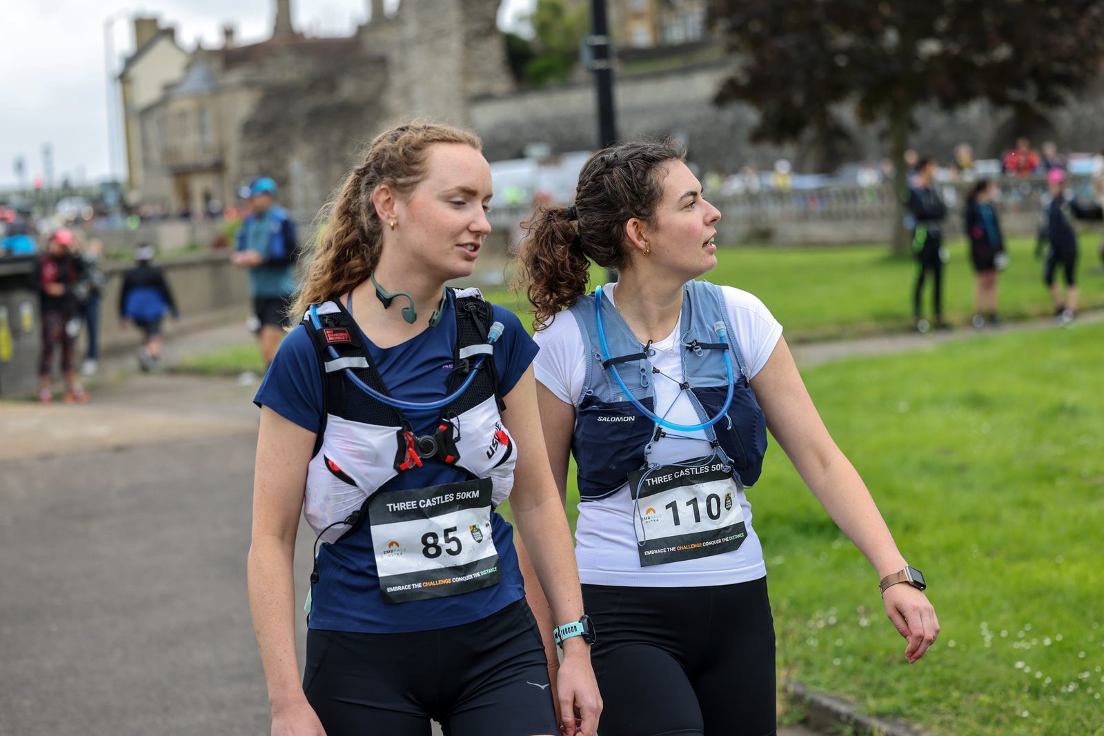 Two women with race bibs, one wearing number 85 and the other 1106, are walking outdoors during a race. They are equipped with hydration packs and wearing athletic gear. Other participants and spectators can be seen in the background near a historic-looking building.