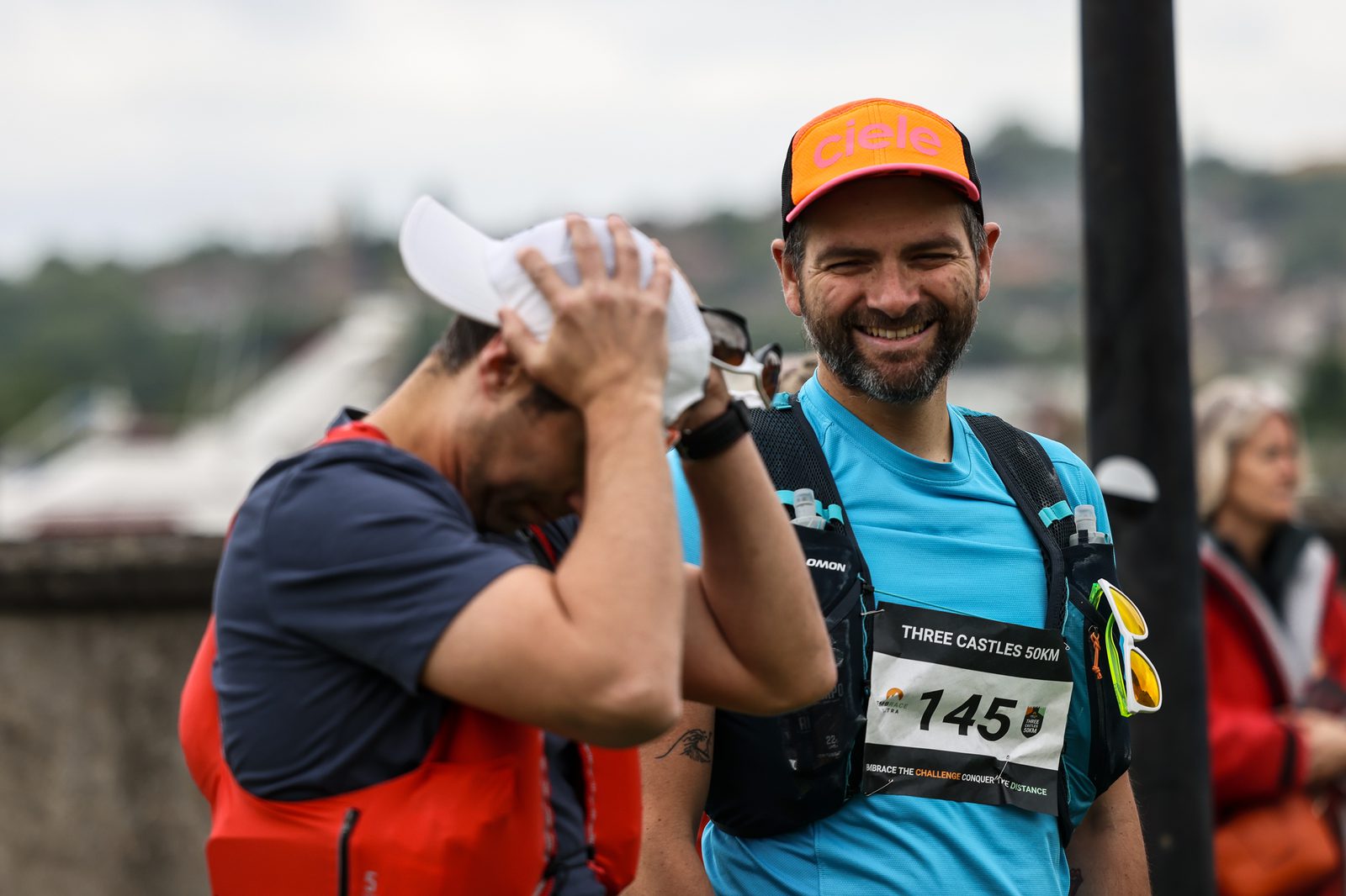 Two men in athletic gear, one wearing a red vest and a white cap, the other in a blue shirt and an orange cap, are preparing for a race. The man in blue, with bib number 145, is smiling, while the other adjusts his cap. Blurred background shows spectators and a cloudy sky.