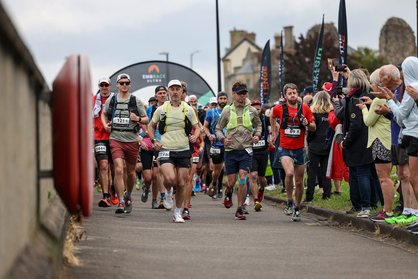 A group of marathon runners, wearing various outfits and numbers, run down a paved road flanked by cheering spectators. They are participating in an outdoor race on a cloudy day. A race banner and flags are visible in the background.