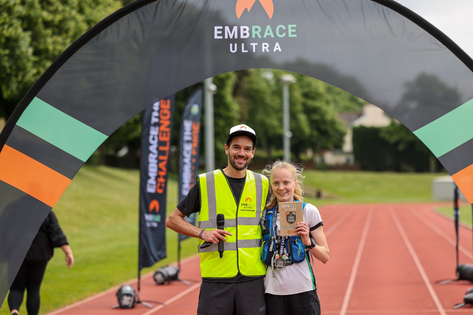 A man and a woman stand together at a running track finish line under an arch that reads "Embrace Ultra." The man in a yellow vest holds a microphone, while the woman, wearing medals, holds a wooden plaque. They both smile at the camera.