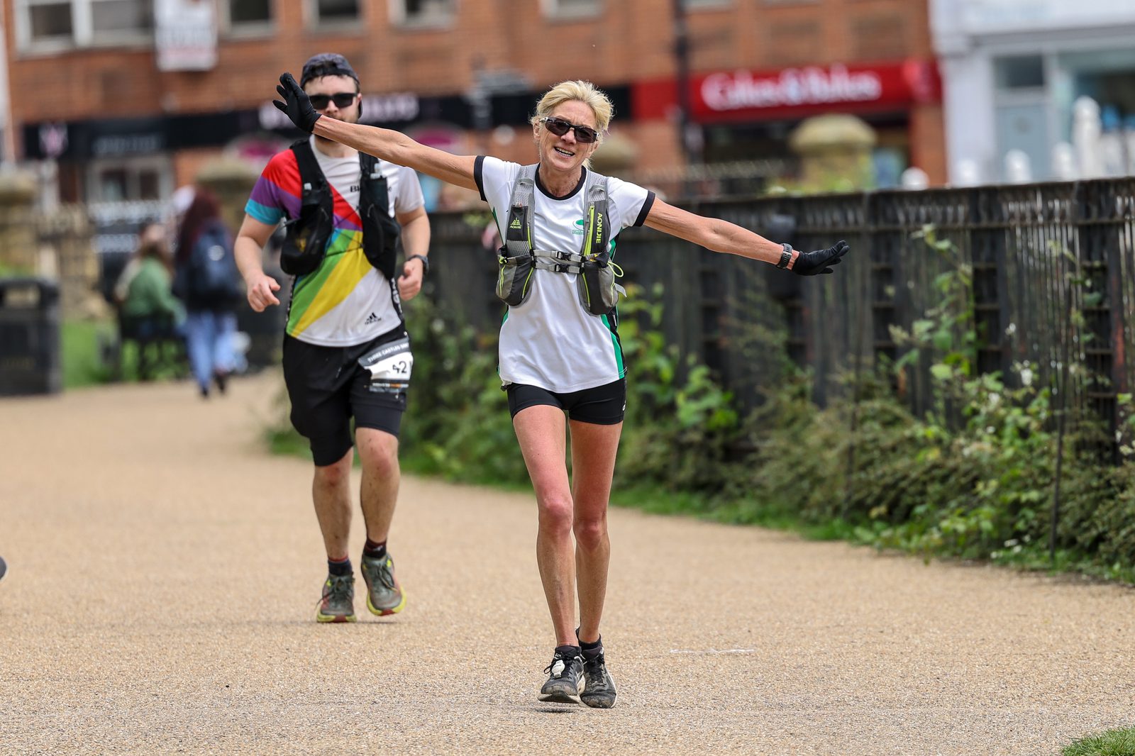A woman dressed in athletic gear runs with her arms outstretched in a joyous manner, while a man runs behind her in similar attire. They are on a pathway outdoors, with greenery and buildings in the background. Both are wearing hydration packs.