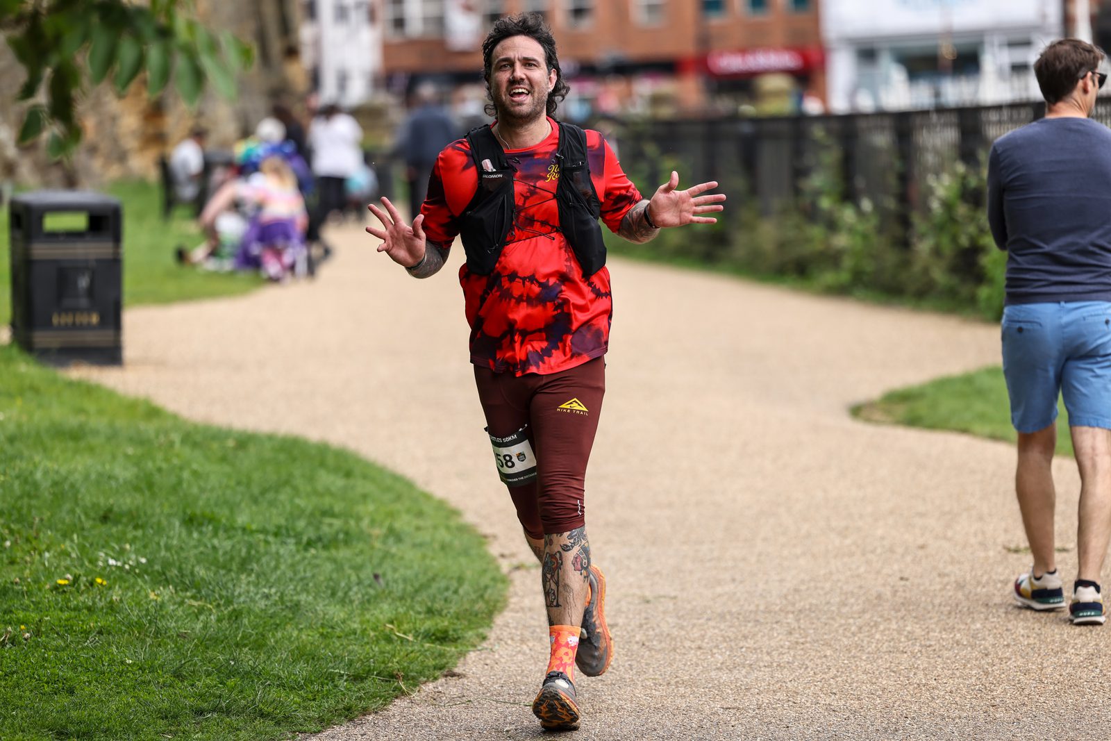 A person wearing a red and black tie-dye shirt and matching shorts with a running number bib joyfully runs on a park path. Other park visitors are seen in the background, enjoying the outdoor space.