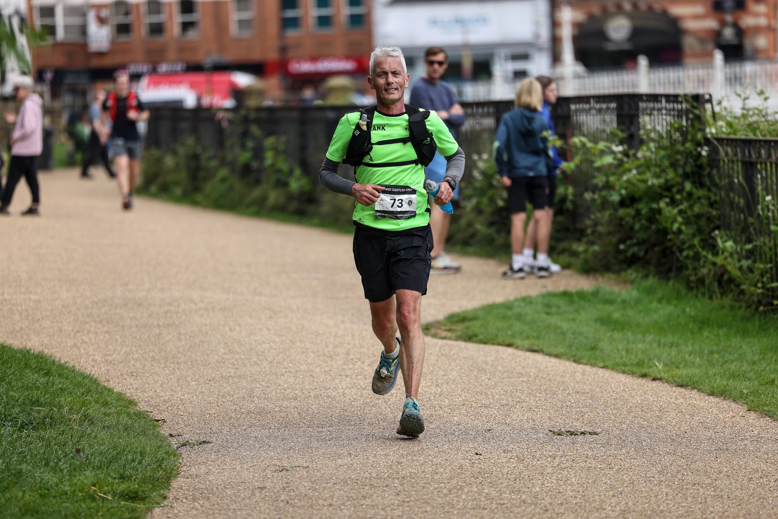A runner wearing a green shirt with the number 734 races down a park pathway. Other runners and spectators are seen in the background. The path is bordered by grass and a fence, and buildings are visible in the distance.