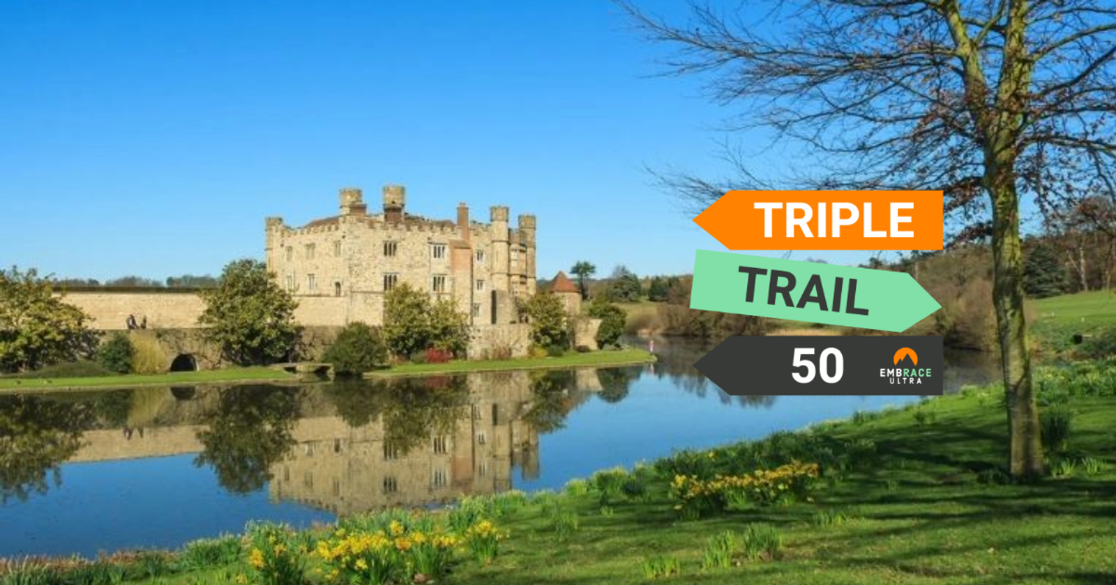 A picturesque castle with multiple turrets stands beside a serene, reflective lake under a clear blue sky. Bright green grass, daffodils, and trees surround the scene. Arrows on the right point to "Triple Trail 50," indicating a hiking or running event.