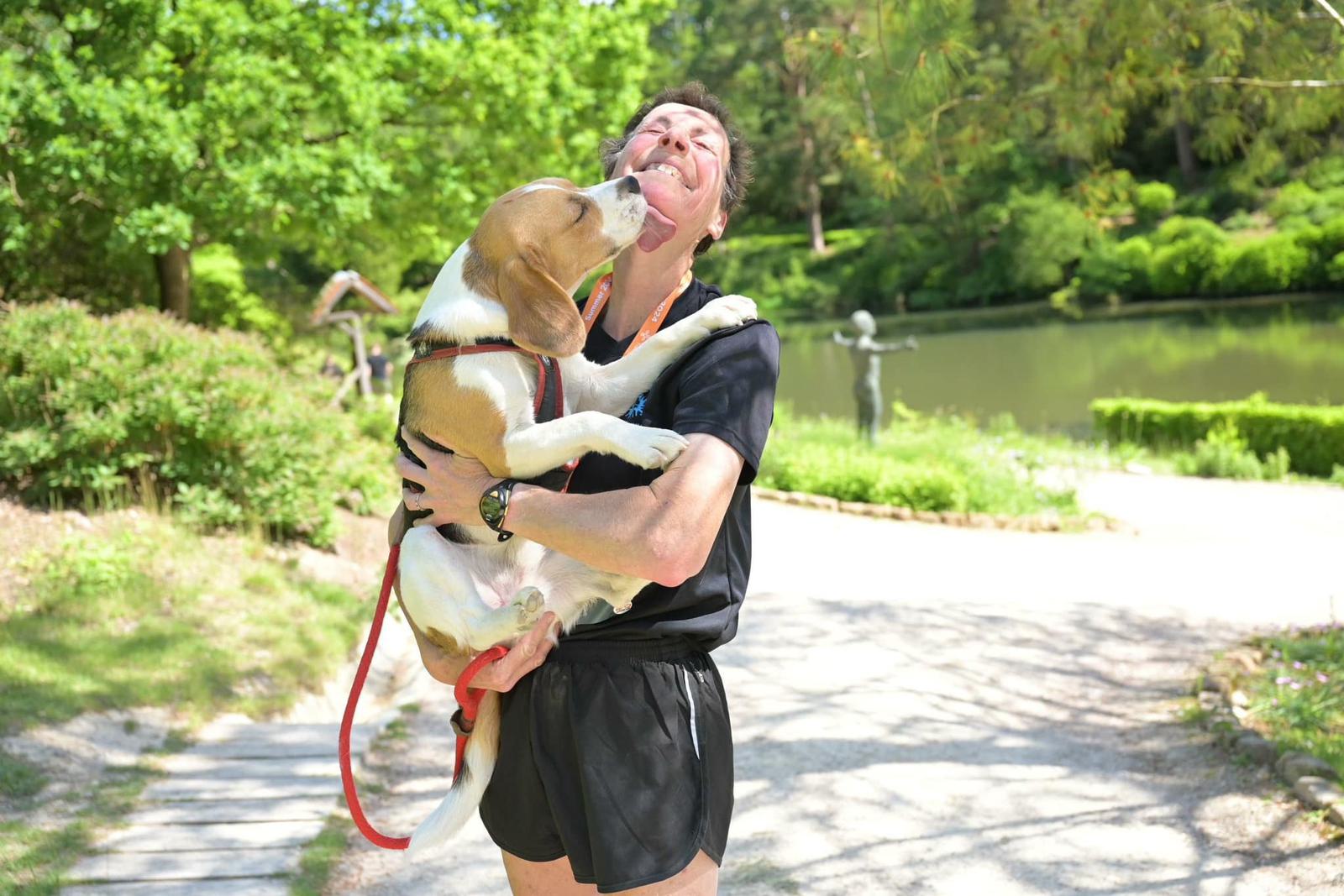 A person in a black outfit holds a beagle dog that is playfully licking their face. They are standing on a path beside a pond, surrounded by lush greenery on a sunny day.