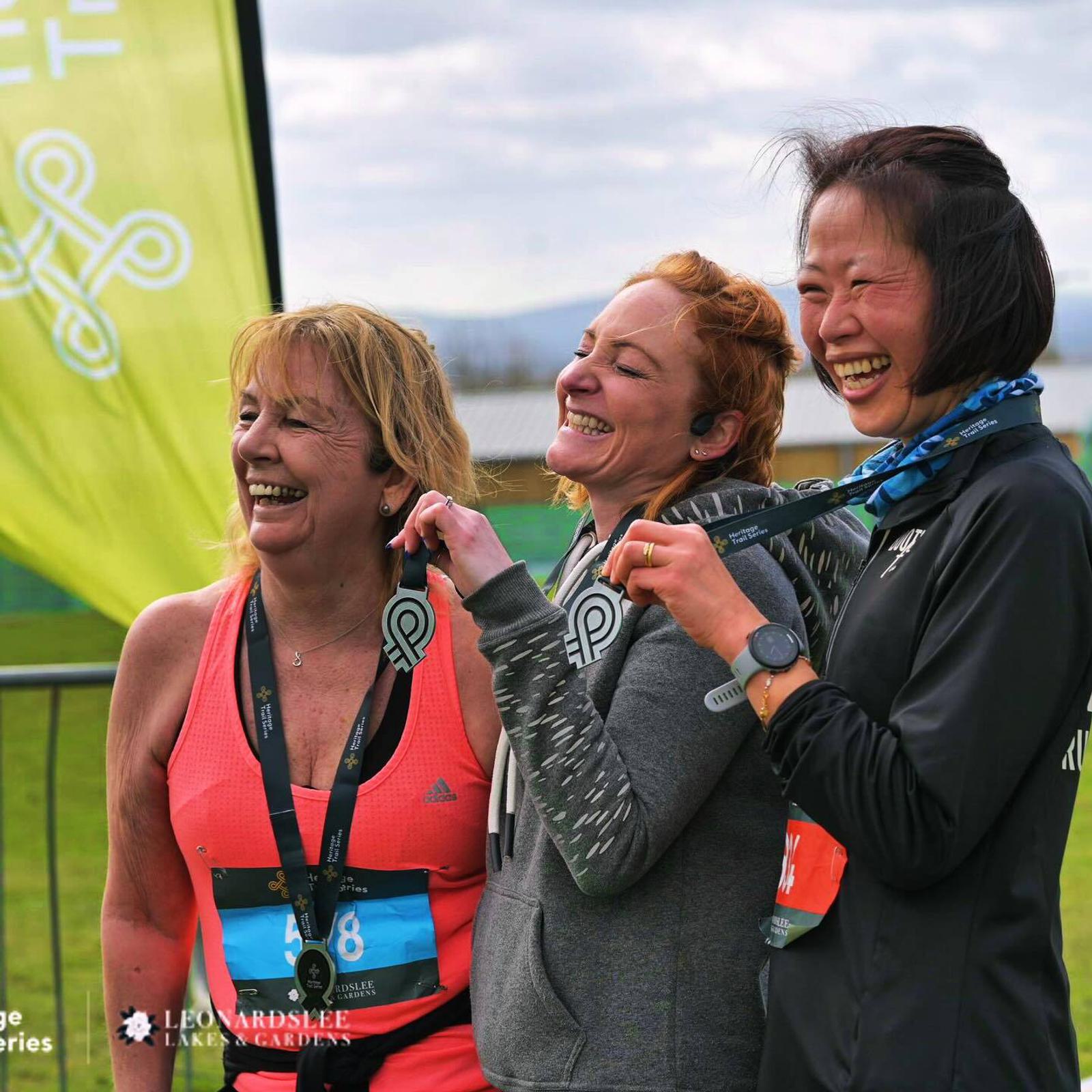 Three smiling women, each holding medals, pose together at a race finish line. They are wearing athletic clothing, and there's a green banner in the background.