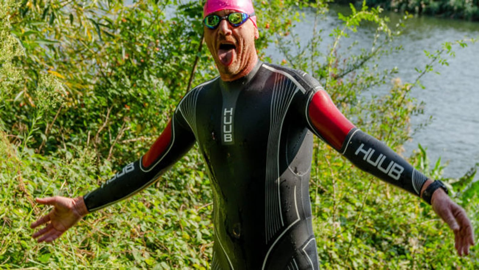 A person wearing a black and red wetsuit and a pink swim cap stands outdoors with arms raised and mouth open, appearing joyful. They are wearing sunglasses and are surrounded by green foliage near a body of water.