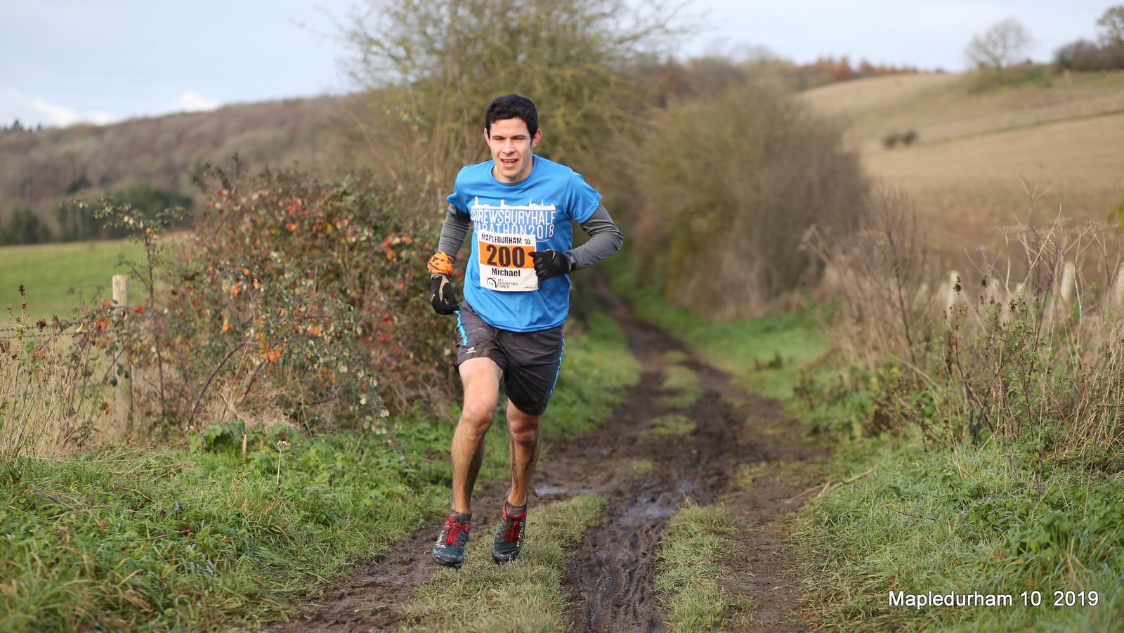 A man wearing a blue shirt and black shorts, running on a muddy trail surrounded by fields and hills. He has a race bib number 200 on his chest. The setting is rural, with some trees and bushes visible. Text on the image reads "Mapledurham 10 2019.