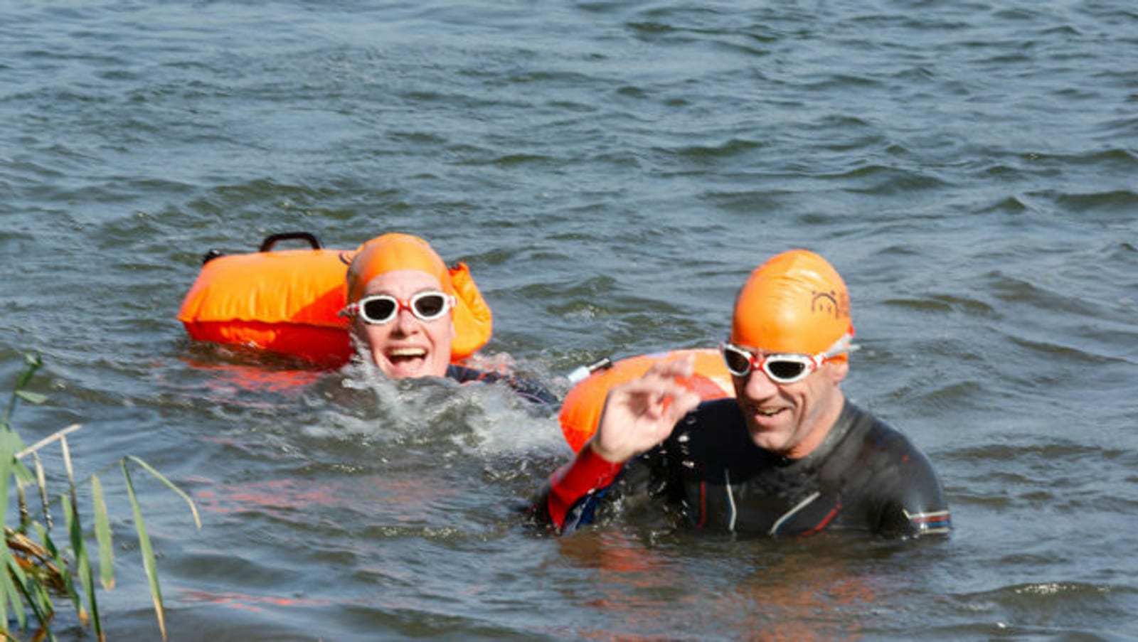 Two smiling swimmers in black wetsuits and orange swim caps float in open water. Each has an orange buoy attached. One swimmer waves at the camera while the other person smiles widely. They both wear white-rimmed goggles, and water plants are visible in the foreground.