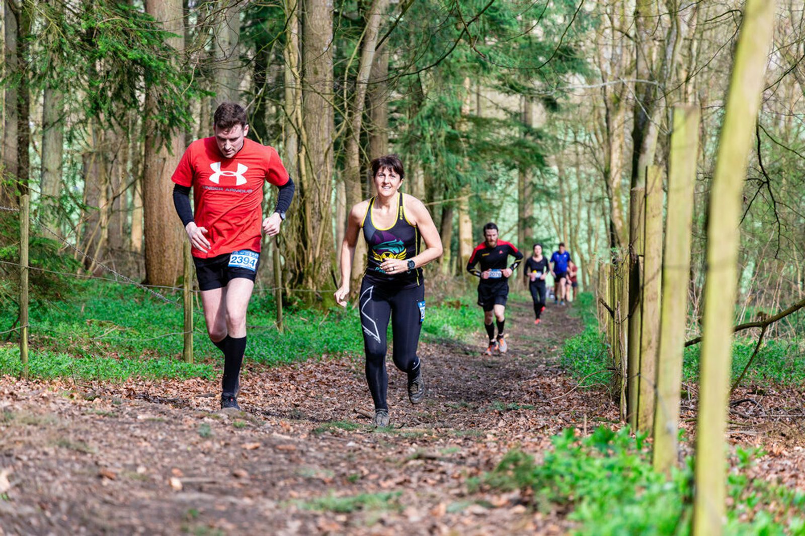 A group of runners navigate a forest trail during a race. In the foreground, a man in a red shirt and a woman in a black tank top are running side by side. Other participants follow behind them on the path, surrounded by green trees and foliage.
