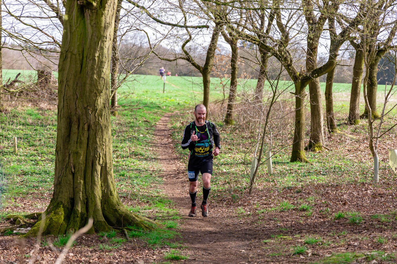 A man is running on a dirt path through a forested area during the day. He is wearing a black athletic outfit with a number on his shorts and making a peace sign with his right hand. The background shows a grassy field with more runners in the distance.