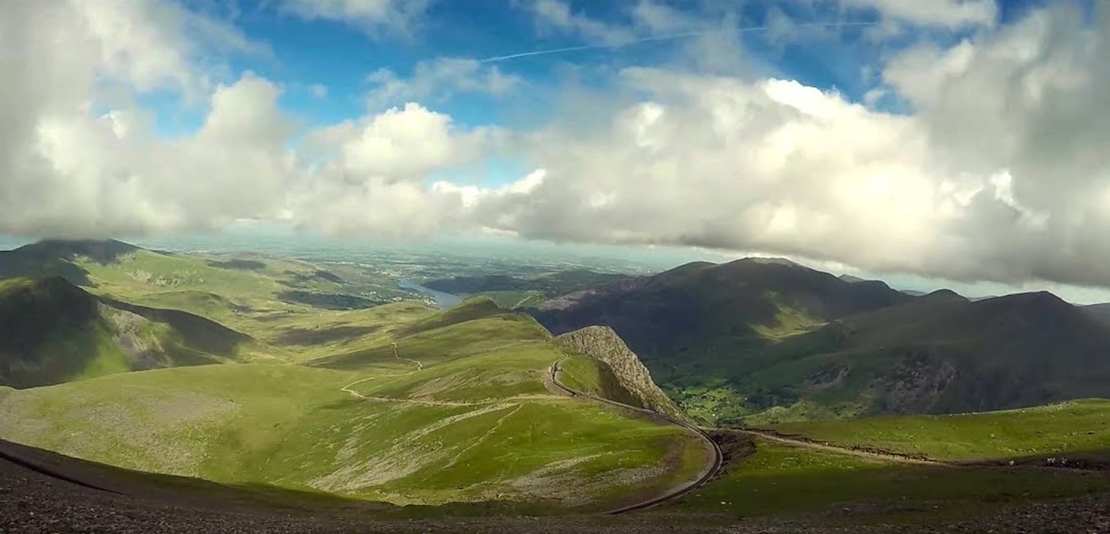 A scenic view of a mountainous landscape under a blue sky with scattered clouds. A winding road cuts through the green and rocky terrain, leading towards distant hills and valleys. The overall scene is serene and expansive.