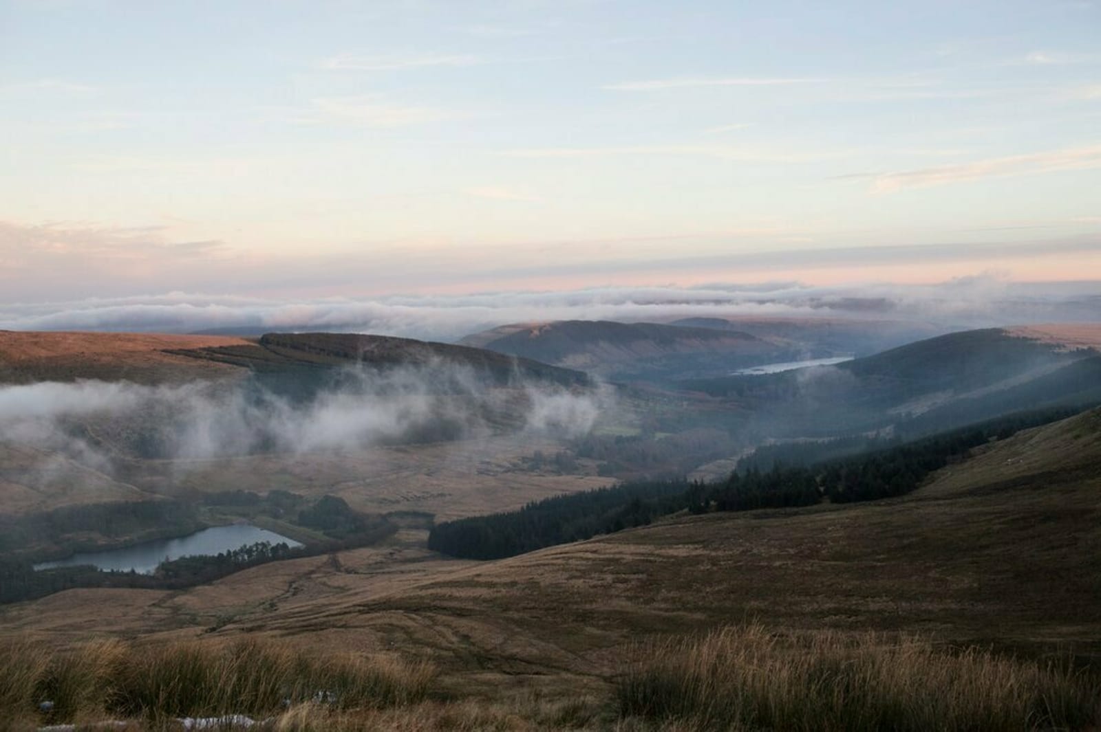 A serene landscape with rolling hills and valleys shrouded in light mist. Low-lying clouds hug the contours of the hills, and a tranquil river meanders through the valley, reflecting the soft light of the early morning or late evening sky.