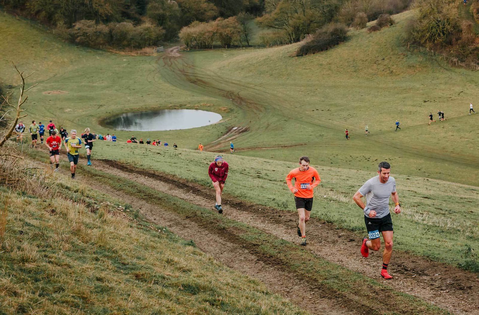 A group of runners participates in a trail race up a grassy hillside. The path is muddy and winds up towards a small pond in the background. The runners are dressed in various colorful athletic outfits. The landscape is dotted with trees and other participants in the distance.