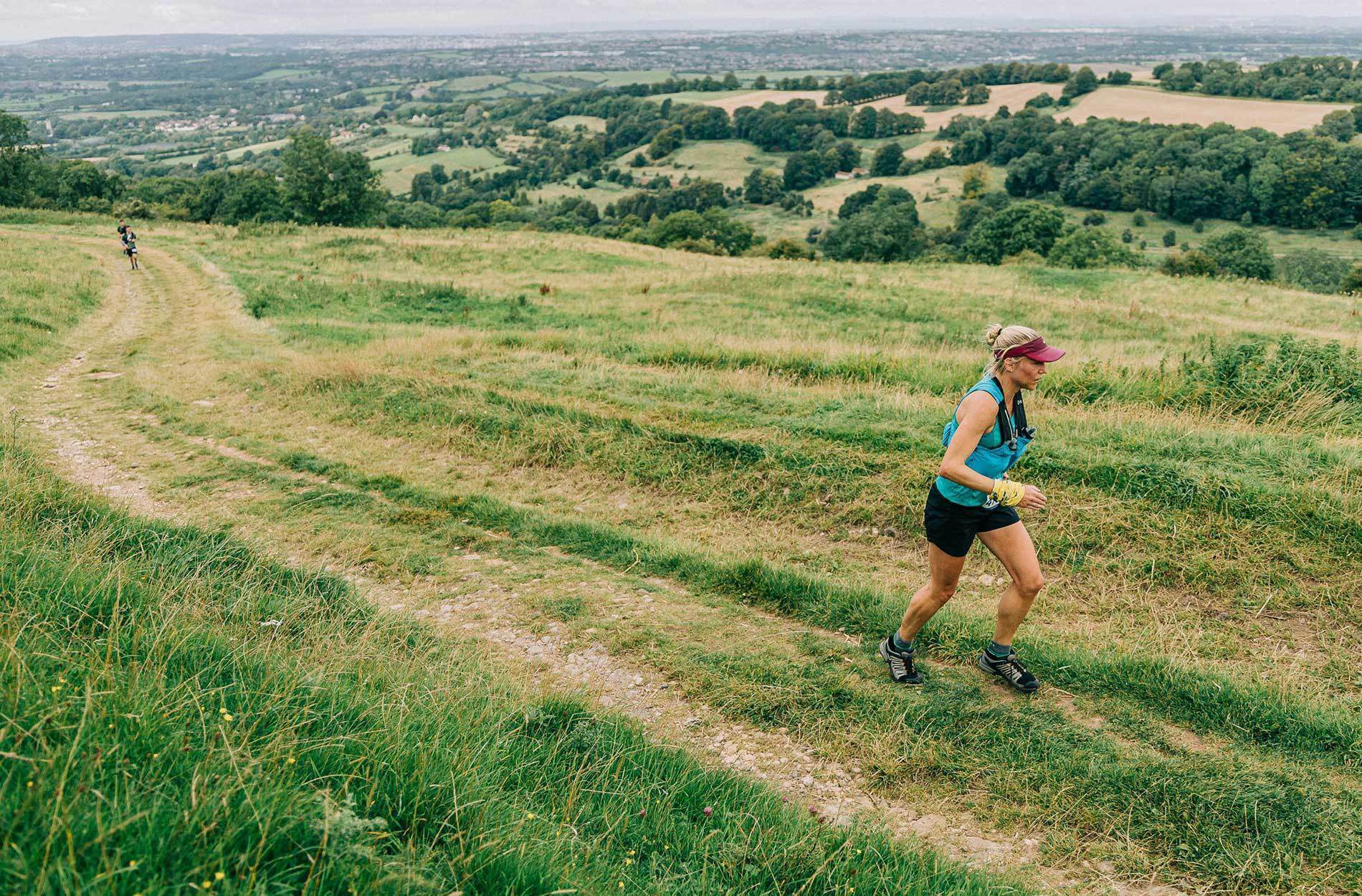 A woman in athletic gear, including a blue tank top, black shorts, and a red visor, runs up a grassy, winding trail on a hillside. Another runner is visible in the distance. The background features scenic rolling hills and a patchwork of fields and trees.