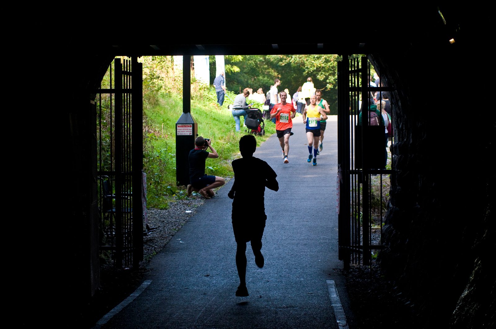 Silhouette of a runner exiting a dark tunnel, with several other runners visible outside in a brightly lit area. Spectators and a photographer are positioned along the sides of the path, capturing the action. The scene depicts a lively outdoor race in progress.