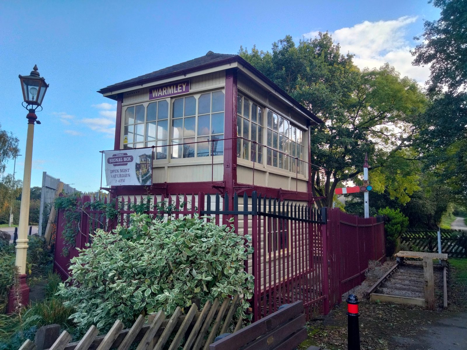 An old railway signal box with "Warmley" on its sign, featuring multiple large windows and burgundy and cream paint. It is surrounded by lush greenery, including trees and a bush with white and green leaves. A lamp post stands on the left, and tracks are visible to the right.