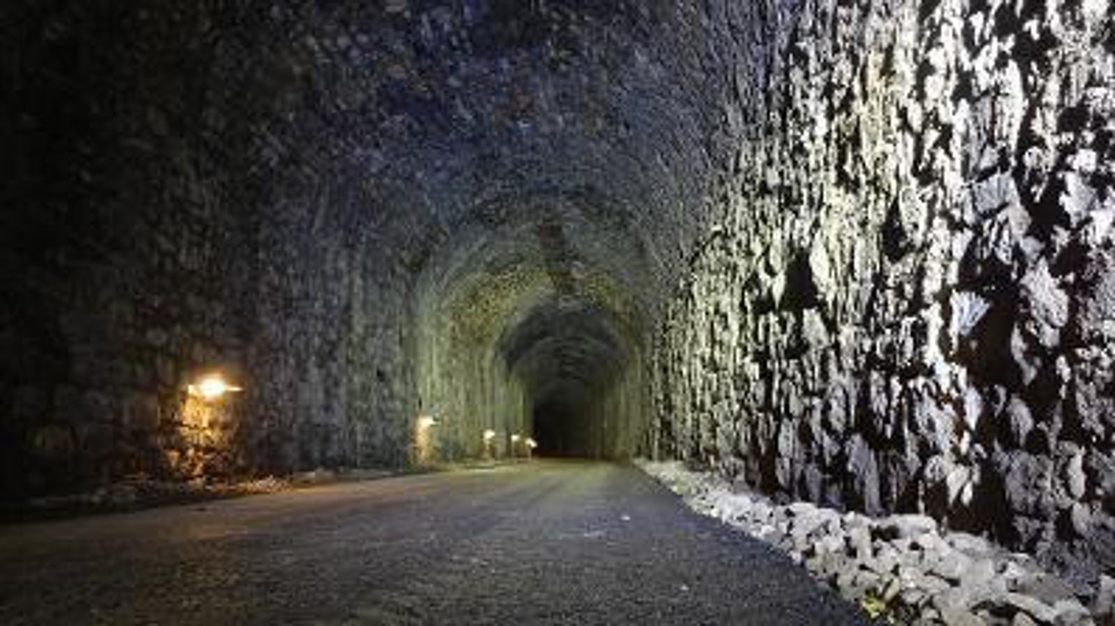 Dimly lit tunnel with rough, textured stone walls. Small lights are mounted intermittently along the left side, casting shadows on the uneven surface. The floor appears to be a gravel pathway extending into darkness. The atmosphere is eerie and serene.