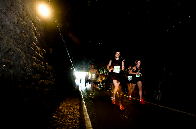 A group of runners jogs through a dimly lit tunnel. The tunnel walls are rugged, and a bright light is visible at the end. The runners wear athletic gear and race bibs, with some visibly smiling as they progress.