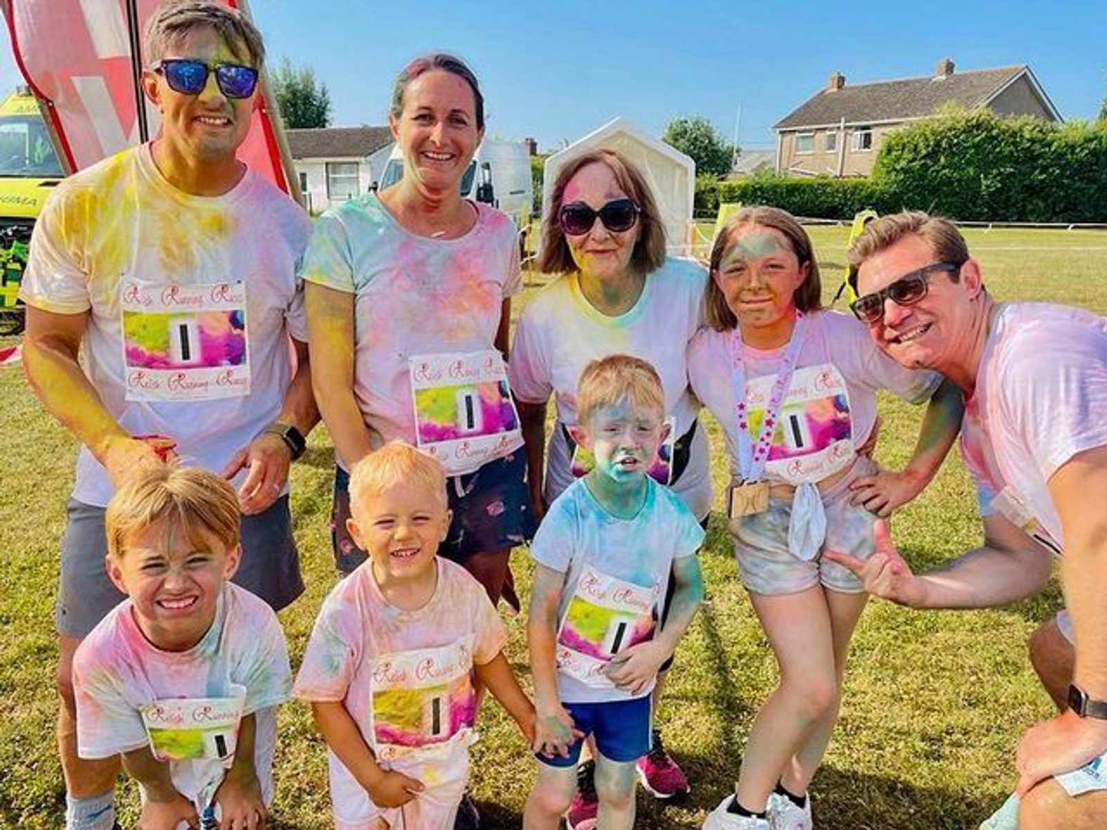 A group of seven people, including three adults and four children, stand in a grassy field, all wearing colorful, powder-splattered clothes and shirts that say "Color Run." They smile at the camera under a bright, sunny sky with houses visible in the background.
