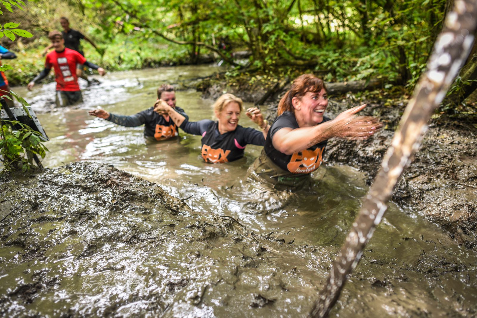 Participants in a mud run navigate through a waterlogged, muddy obstacle in a forested area. They appear to be enjoying the challenging activity, smiling and helping each other through the thick mud. Green foliage surrounds the scene, and sunlight filters through the trees.