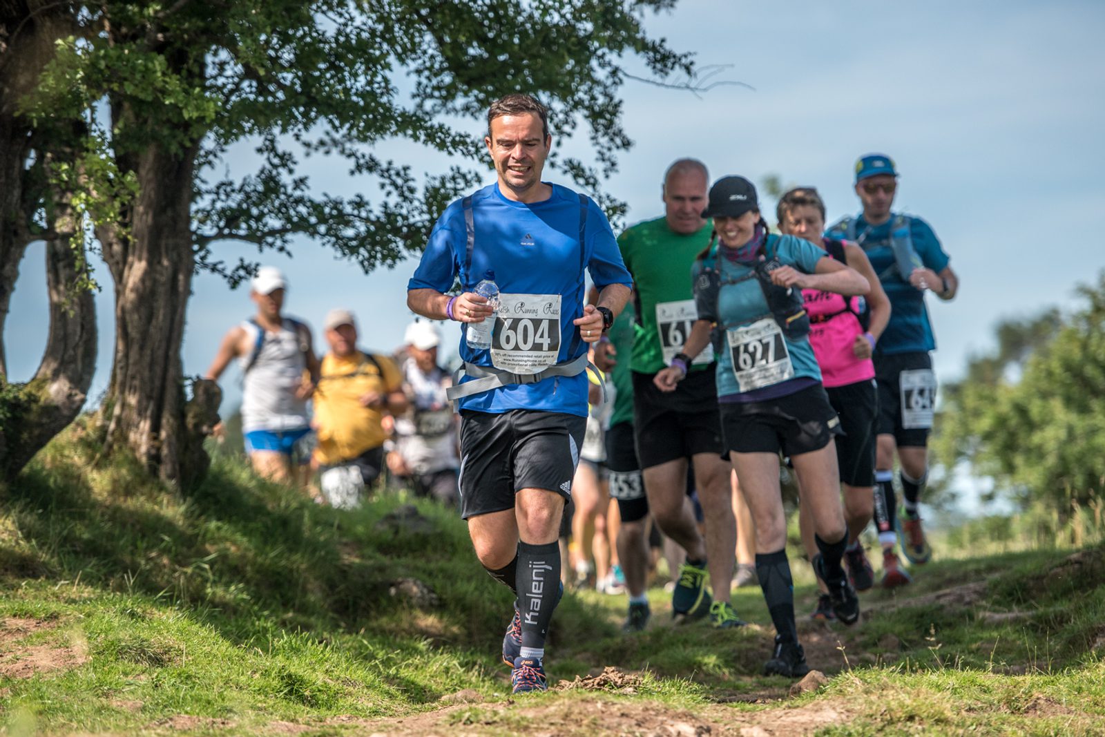 A group of runners, wearing race numbers, are participating in a trail run on a sunny day. The leading runner, in a blue shirt and black shorts, is smiling. They are running on a grassy, uneven path with trees on the left and blue skies above.