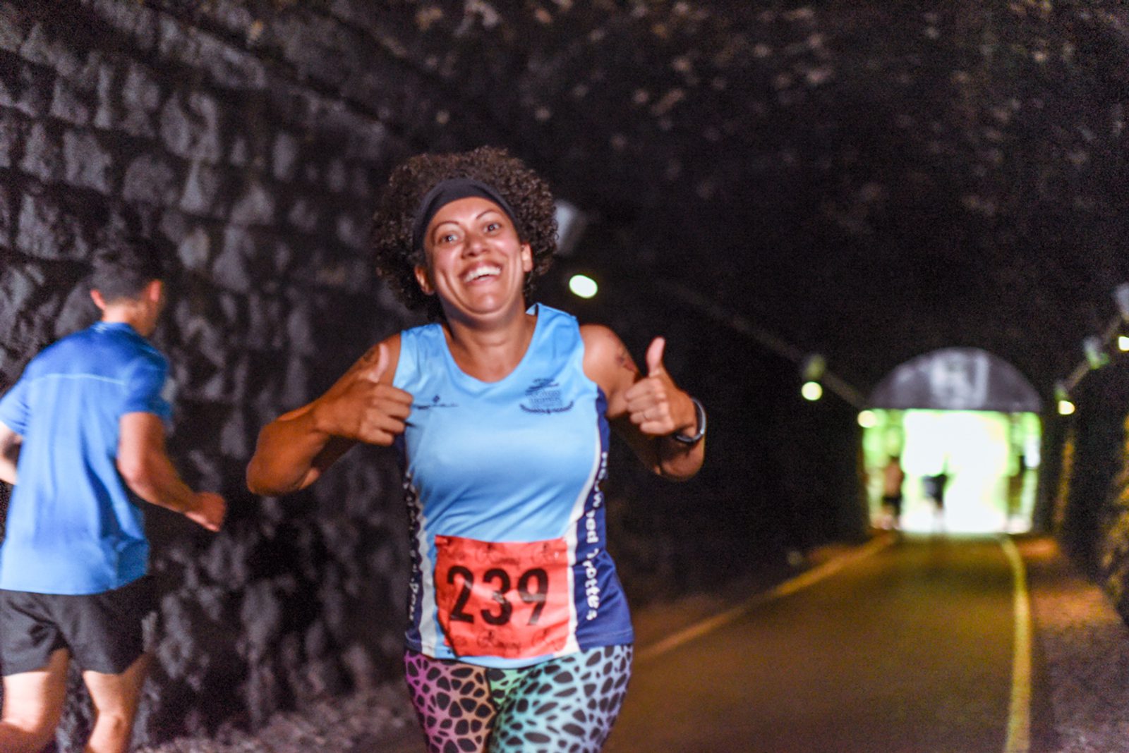 A woman wearing a blue tank top, colorful patterned leggings, and a race bib number 233 gives two thumbs up as she runs inside a dimly lit tunnel. Another runner is visible in the background, and bright light can be seen at the tunnel's exit.