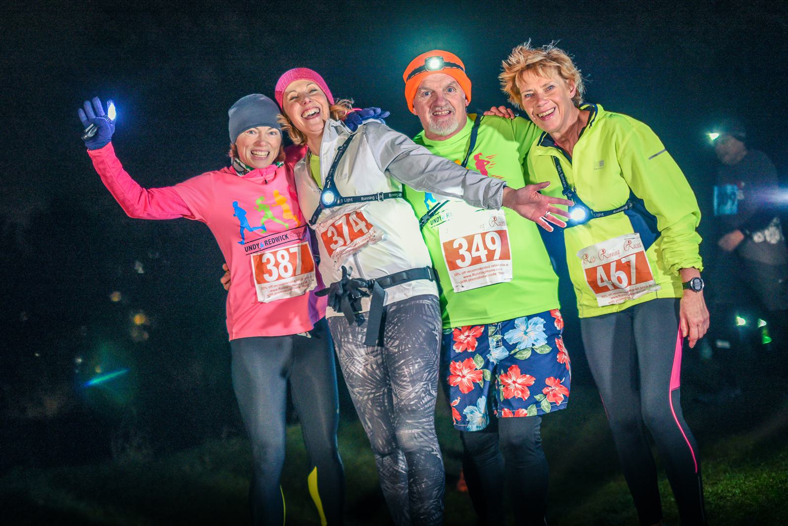 Four runners wearing numbered bibs pose and smile for a photo in the dark. They are dressed in colorful athletic gear, including headlamps and gloves. The background is dimly lit, suggesting a nighttime race or event.