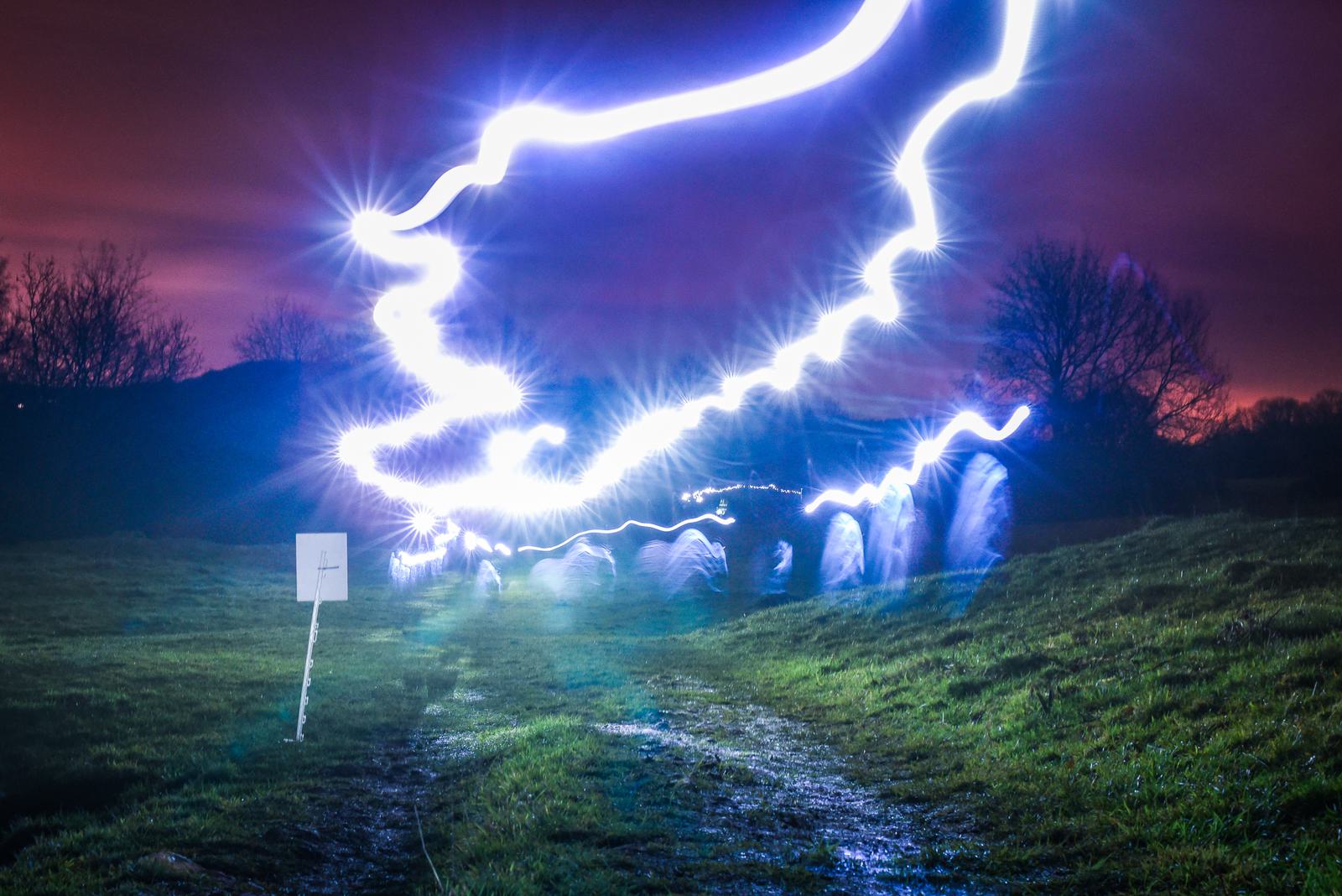 A night scene with swirling blue light trails above a field. The sky is a mix of purple and orange hues, and there are silhouetted trees in the background. A small white sign is visible on the left side of the field.