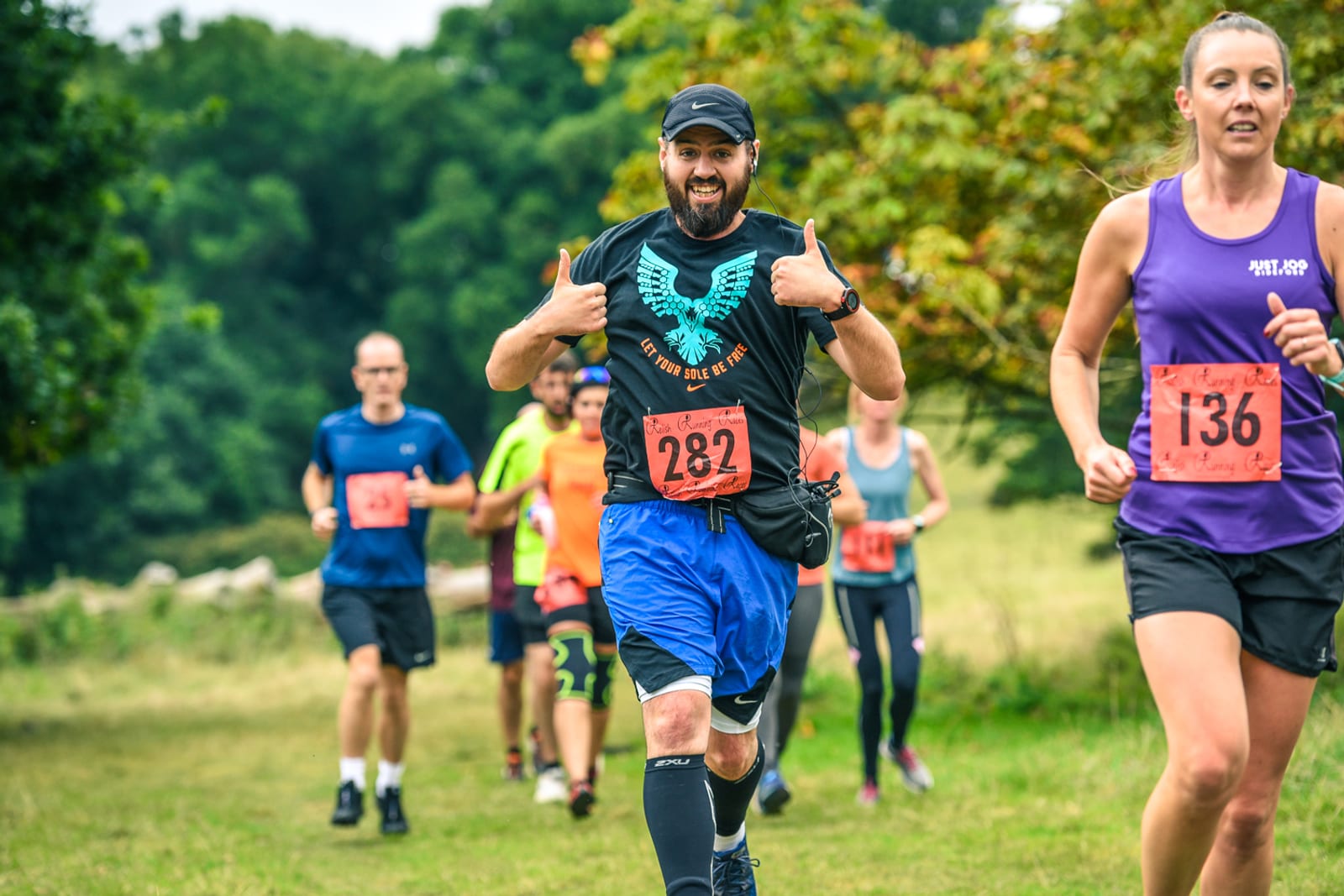 A group of runners participating in a race on a grassy field. The man in the foreground, wearing a black cap, black shirt, blue shorts, and race bib 282, is smiling and giving two thumbs up. Other runners, including a woman in a purple top, are visible in the background.