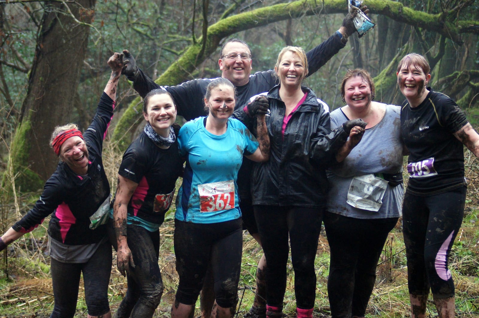 A group of seven people, smiling and covered in mud, pose together in a wooded area. They appear to have participated in an outdoor, possibly muddy, event or obstacle course. Several members wear numbered bibs on their outfits. The trees and ground are damp.