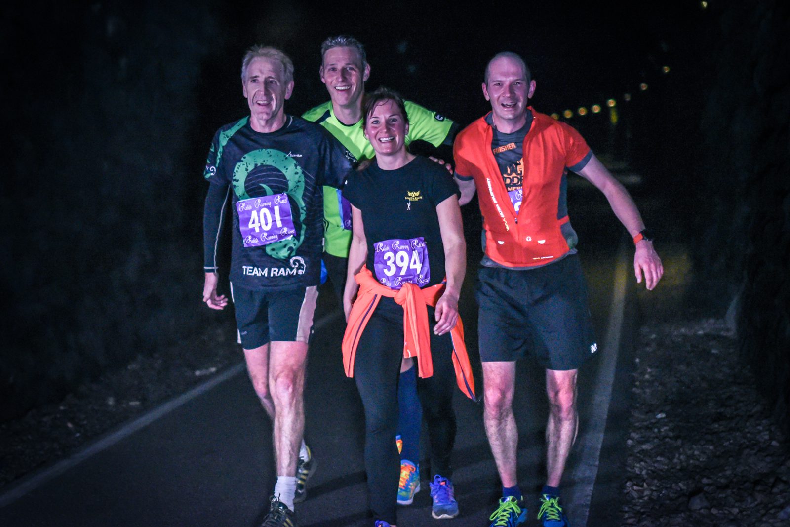 Four runners, three men and one woman, are smiling as they jog or walk together along a dimly lit path at night. They are wearing race bibs numbered 401 and 394. The group appears to be enjoying the night run, embracing camaraderie and teamwork.
