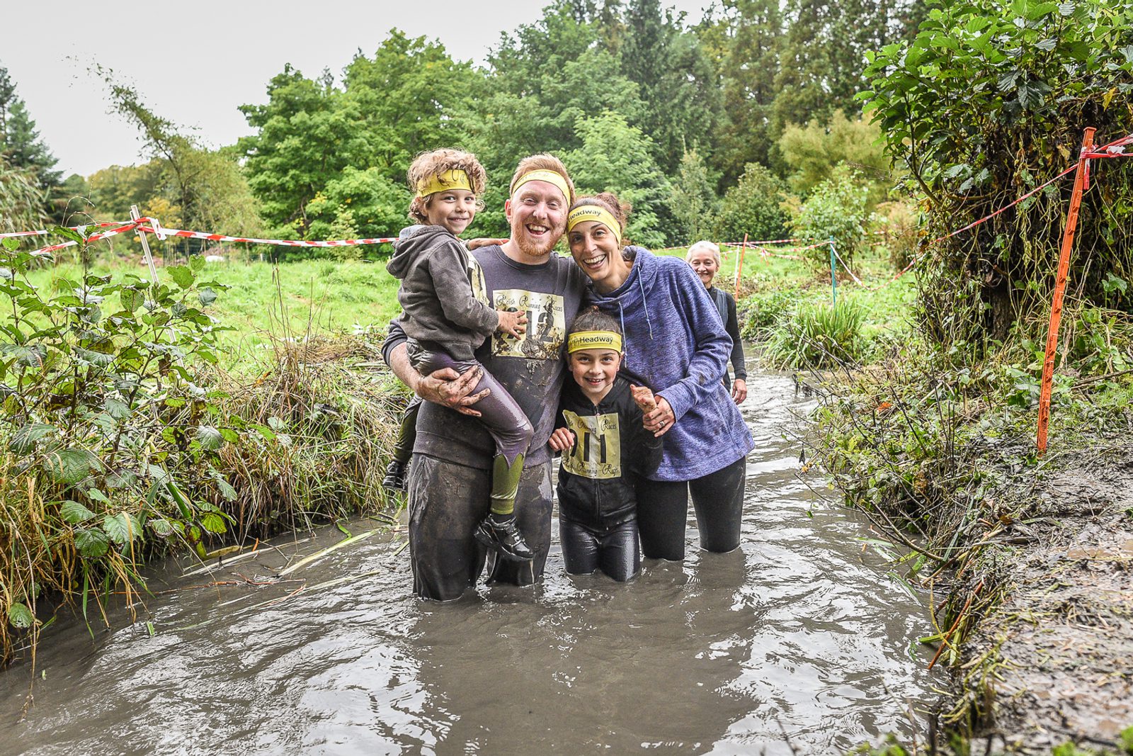A family of four stands in a muddy stream, smiling and posing for the camera. They are participating in an outdoor adventure or obstacle course event, wearing matching headbands and sports attire. The background shows a lush, green wooded area with some terrain markers.