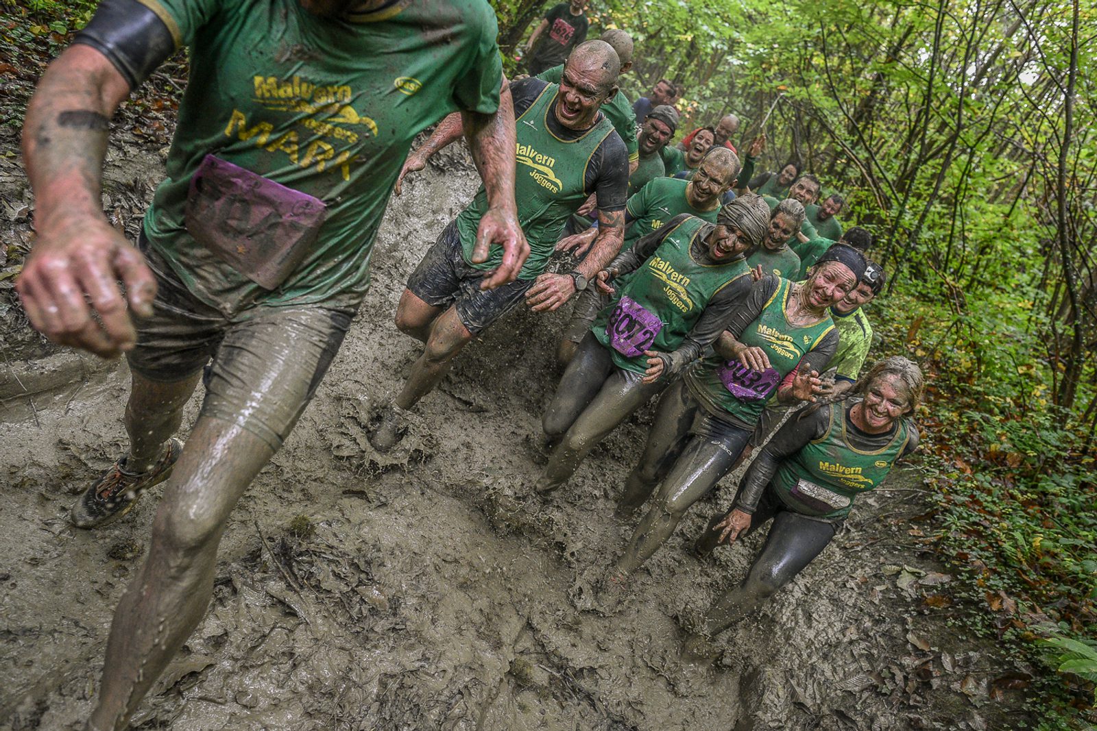A group of people wearing green shirts and race bibs run through a muddy forest trail, covered in mud from head to toe. They appear to be participating in an obstacle course or adventure race, smiling and laughing despite the challenging conditions.