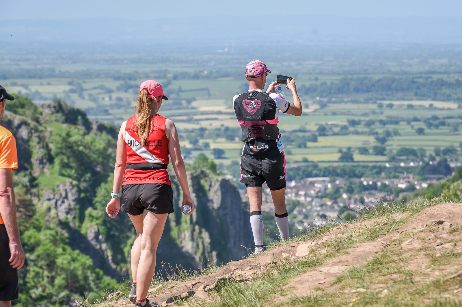 Two runners are on a trail overlooking a scenic valley. One, wearing a red shirt, walks while the other, in a black and pink outfit, stops to take a photo with a smartphone. The landscape features rolling green fields and distant mountains under a clear sky.