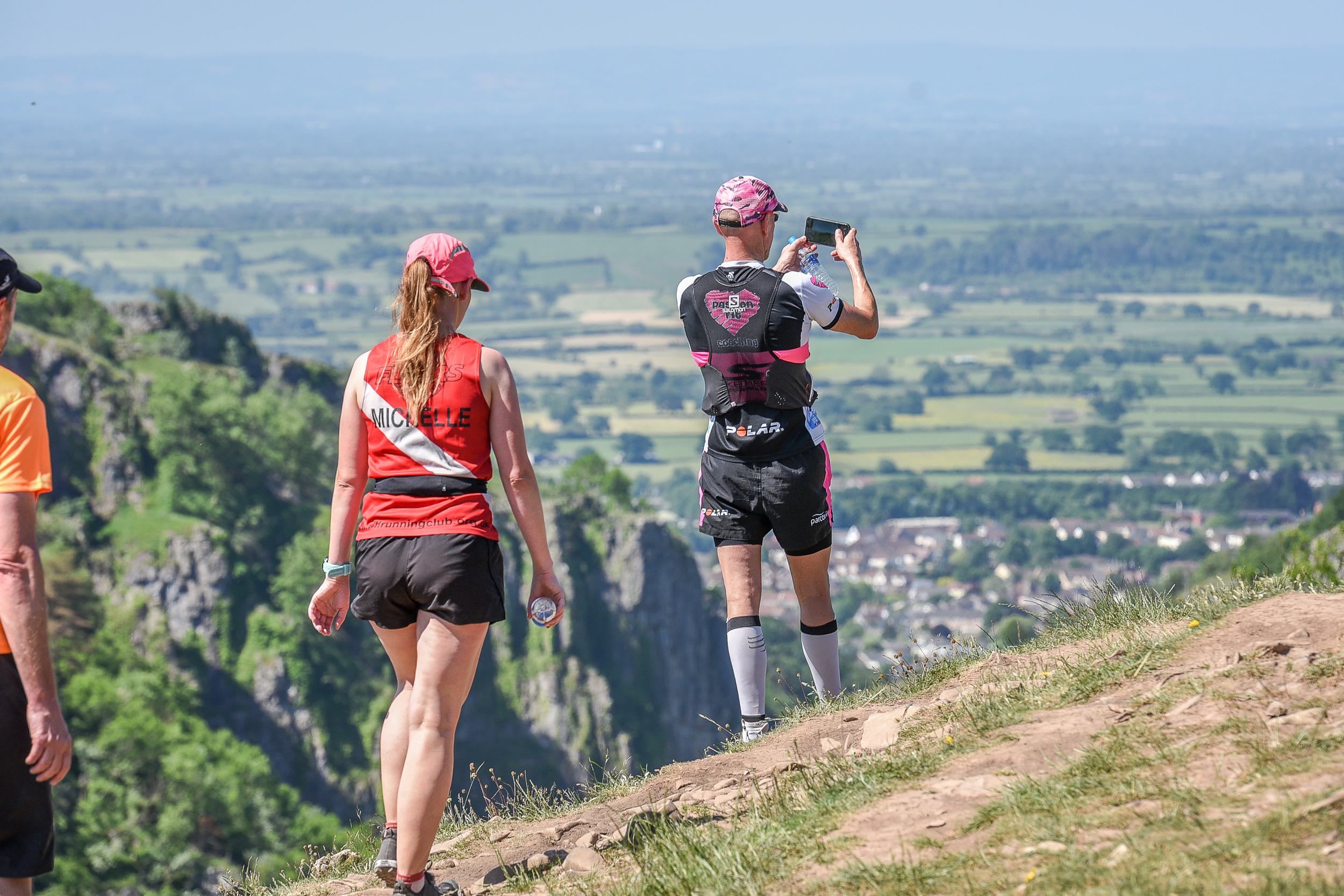 Two runners are on a trail overlooking a scenic valley. One, wearing a red shirt, walks while the other, in a black and pink outfit, stops to take a photo with a smartphone. The landscape features rolling green fields and distant mountains under a clear sky.
