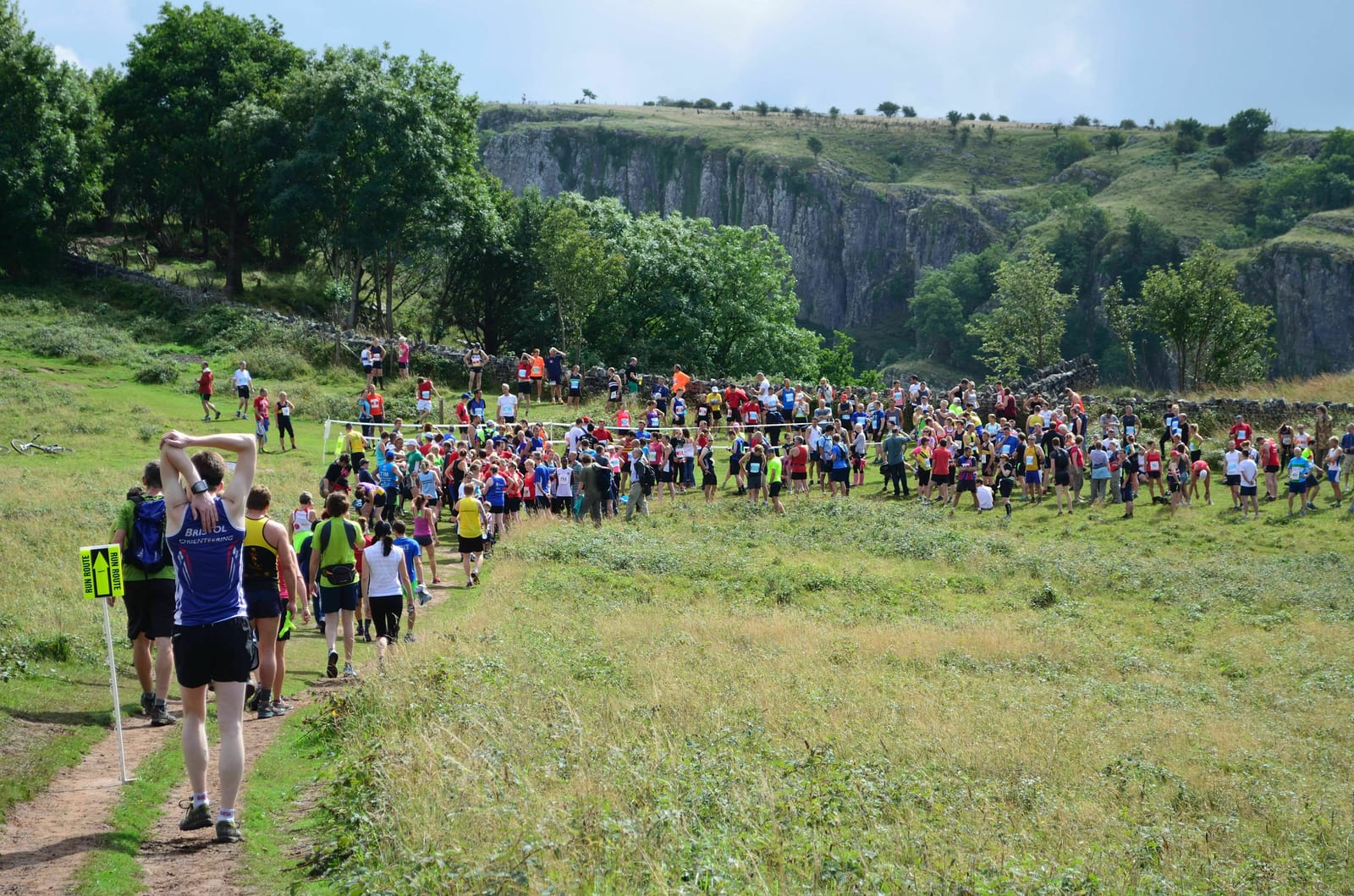 A large group of runners participating in a trail race, making their way through lush green fields and hills. The landscape features a cliffside in the background and various trees. Some runners are wearing colorful outfits, with one holding a staff in the foreground.