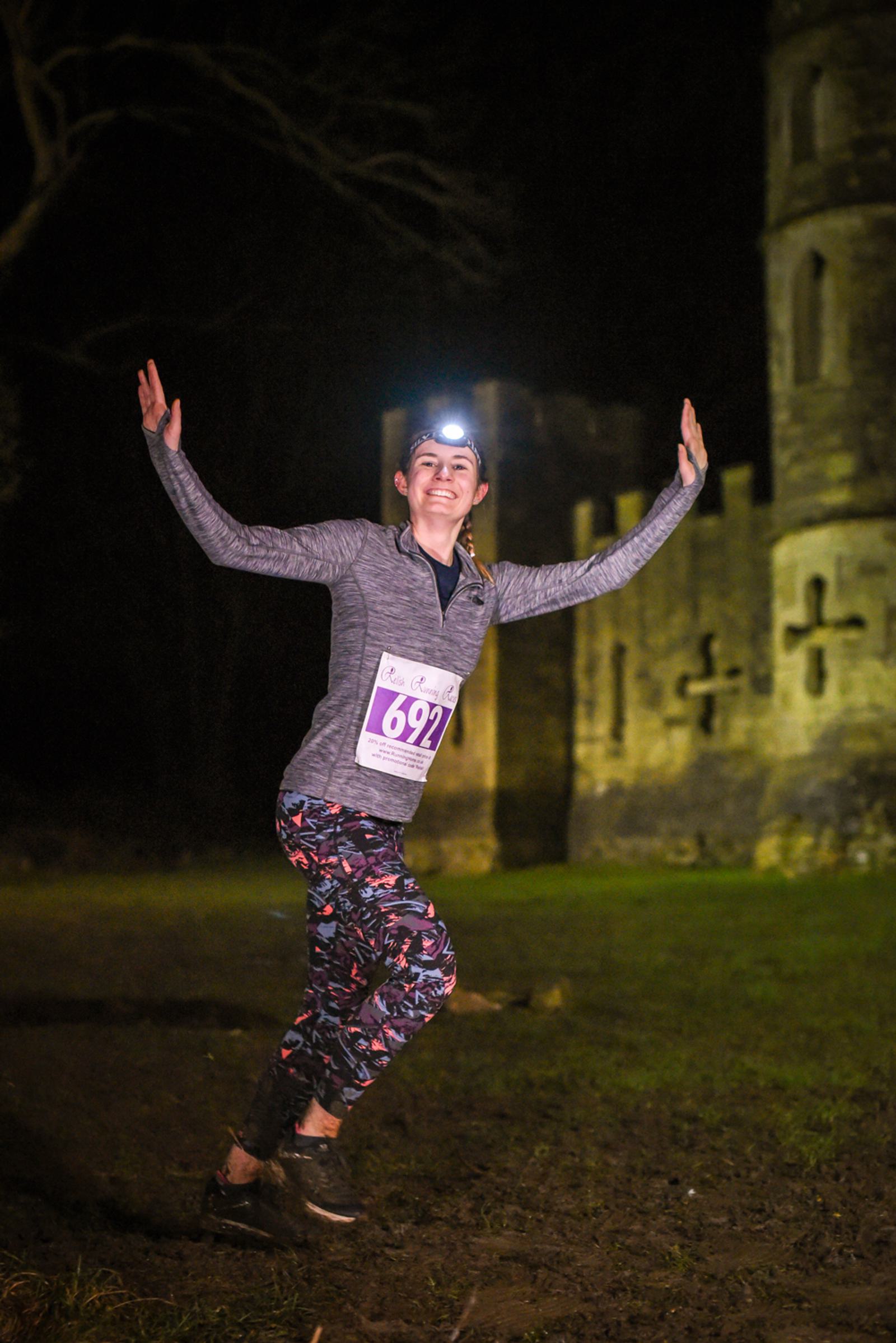 A person wearing a headlamp and bib number 692 is joyfully posing while running at night. They are in front of an old stone building, possibly a castle. The person is dressed in a long-sleeve shirt and patterned leggings.