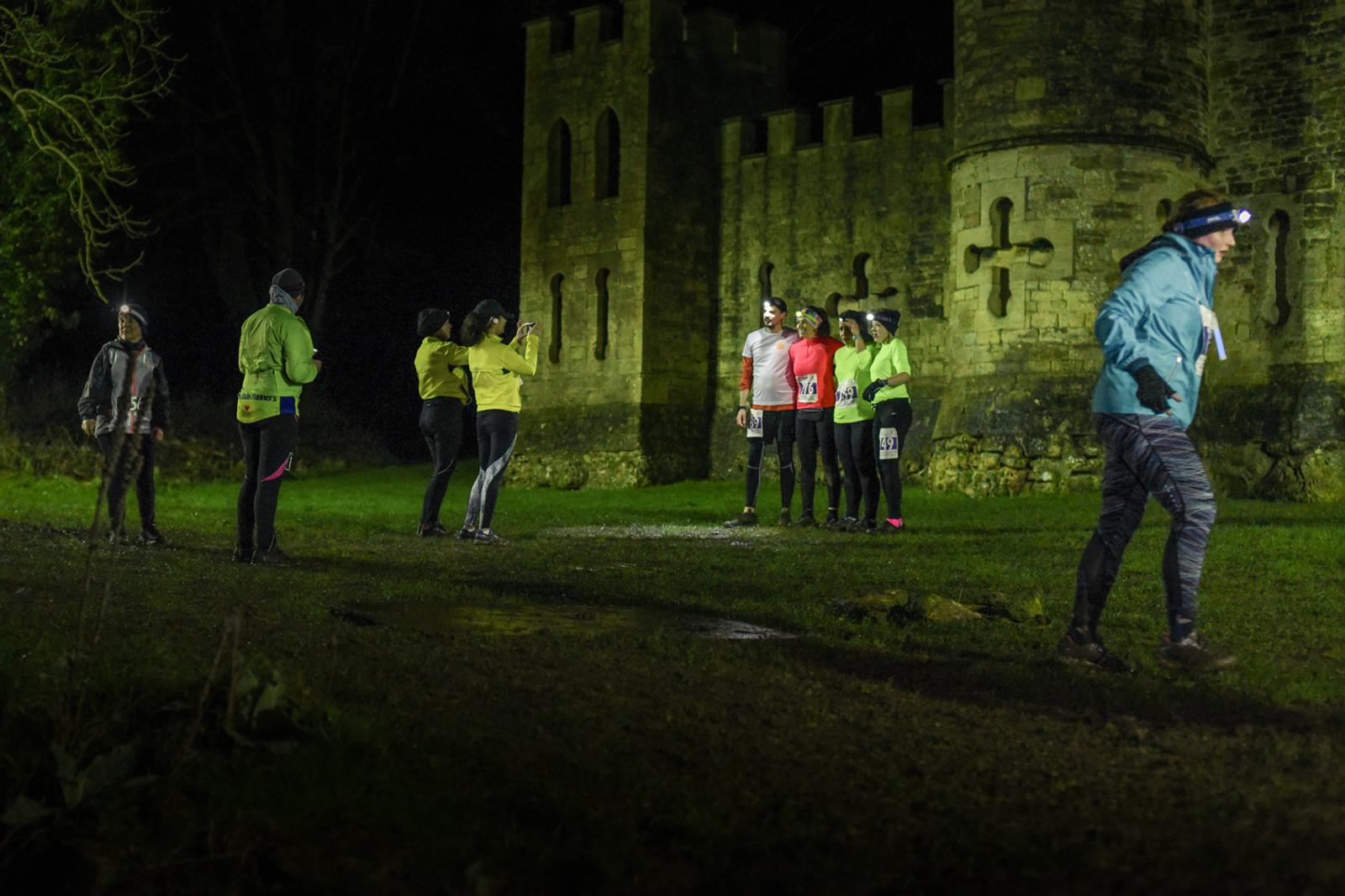 A group of people wearing running gear and headlamps stand and chat near a castle at night. The illuminated building provides a dramatic backdrop as a few individuals pose for a photo. Two people walk away to the right.