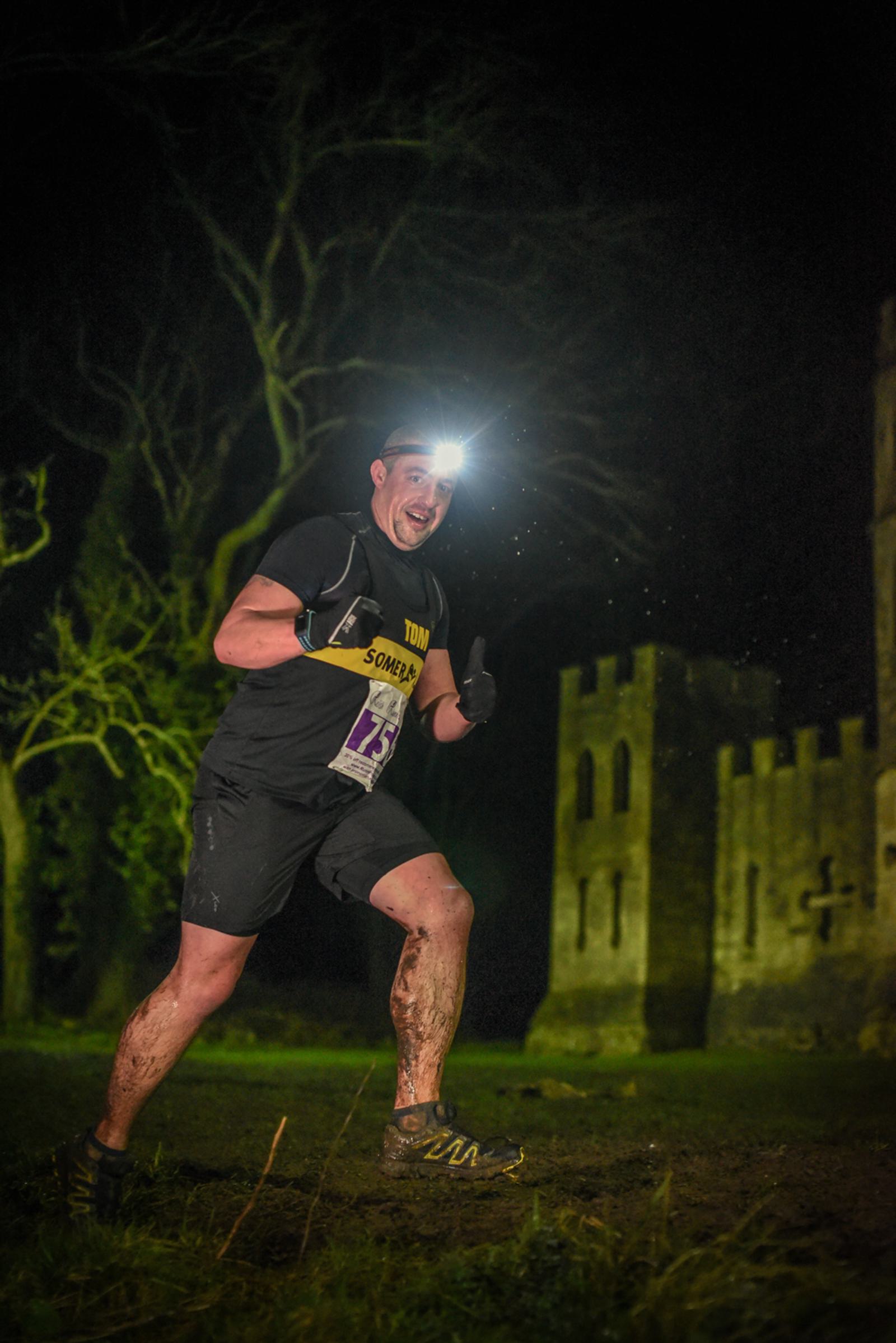A runner wearing a headlamp runs energetically at night past a castle-like structure. The person is smiling and giving a thumbs-up gesture, covered in mud and dressed in a race bib and athletic gear. Trees and a tower are visible in the dark background.