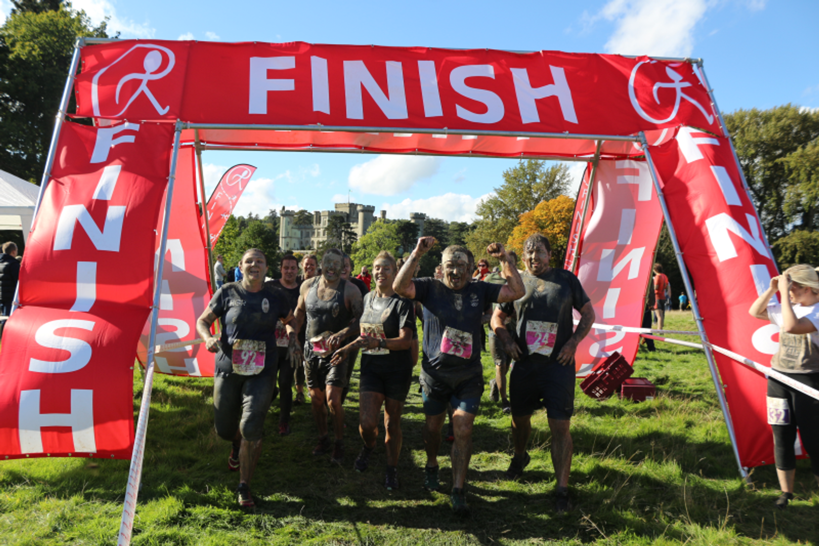 A group of muddy and smiling runners cross the finish line of an outdoor race under a red "FINISH" banner. They appear excited and victorious, holding hands and raising arms in celebration. The background includes trees and the clear sky overhead.
