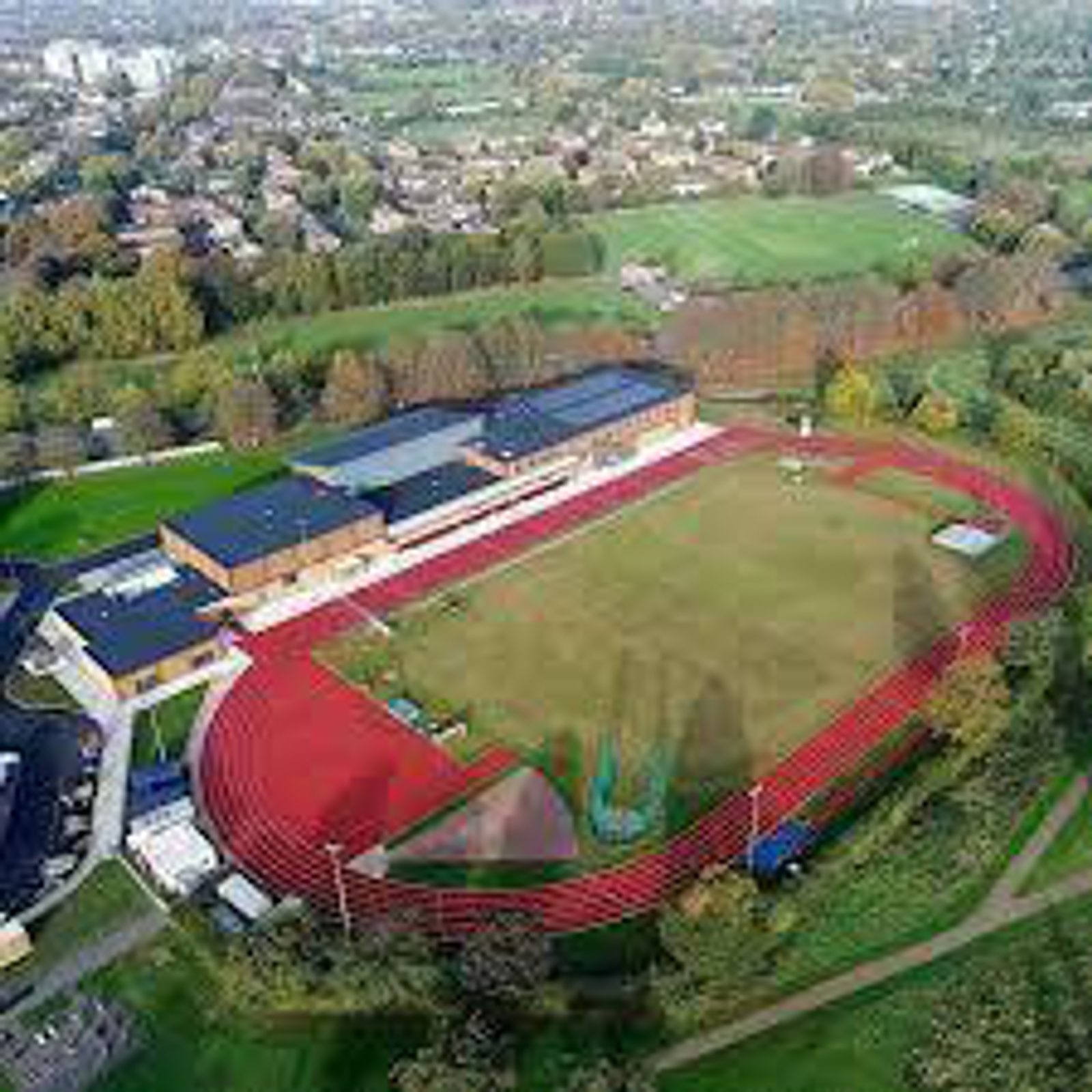 An aerial view of an outdoor sports complex featuring a red running track surrounding a green field, a cluster of buildings next to the track, and surrounding greenery. Residential areas and additional green spaces are visible in the background.
