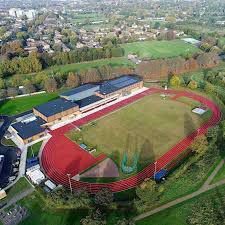 An aerial view of an outdoor sports complex featuring a red running track surrounding a green field, a cluster of buildings next to the track, and surrounding greenery. Residential areas and additional green spaces are visible in the background.