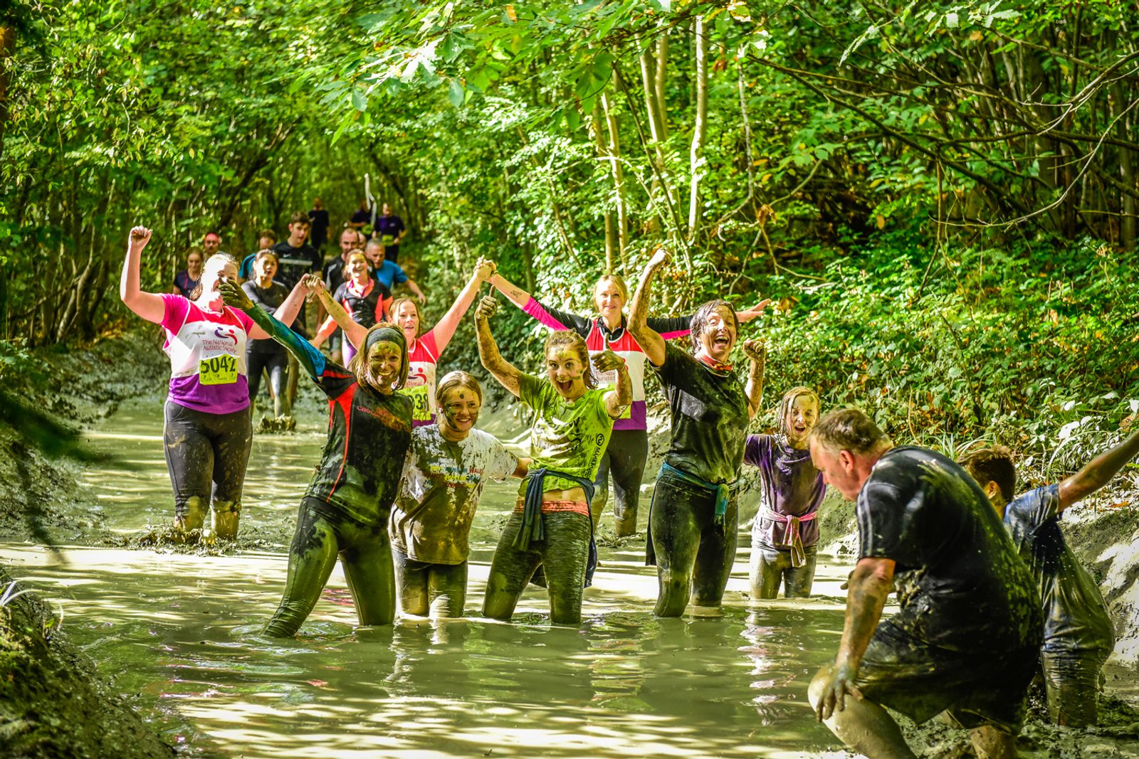 A group of people, covered in mud, joyfully raise their arms as they navigate a muddy, forest trail during a race. The runners in the background are also muddy and smiling, surrounded by dense green trees under bright sunlight. Race bibs are visible on some participants.