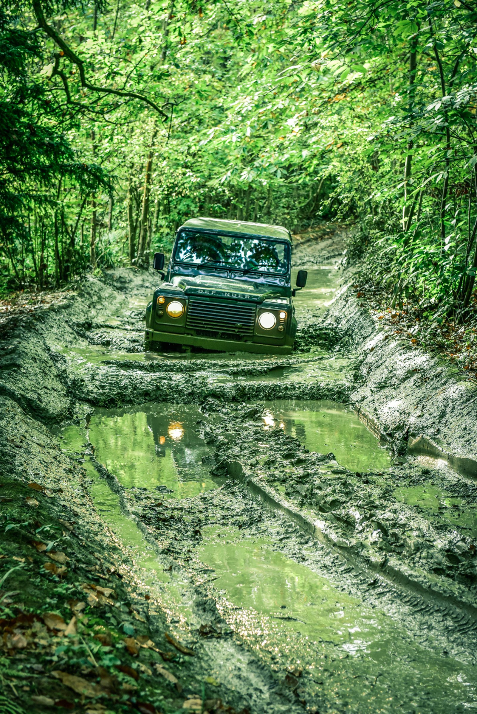 A green off-road vehicle drives through a muddy, narrow forest path filled with deep puddles, surrounded by lush green foliage and trees. The vehicle's headlights are on, illuminating the challenging terrain ahead.