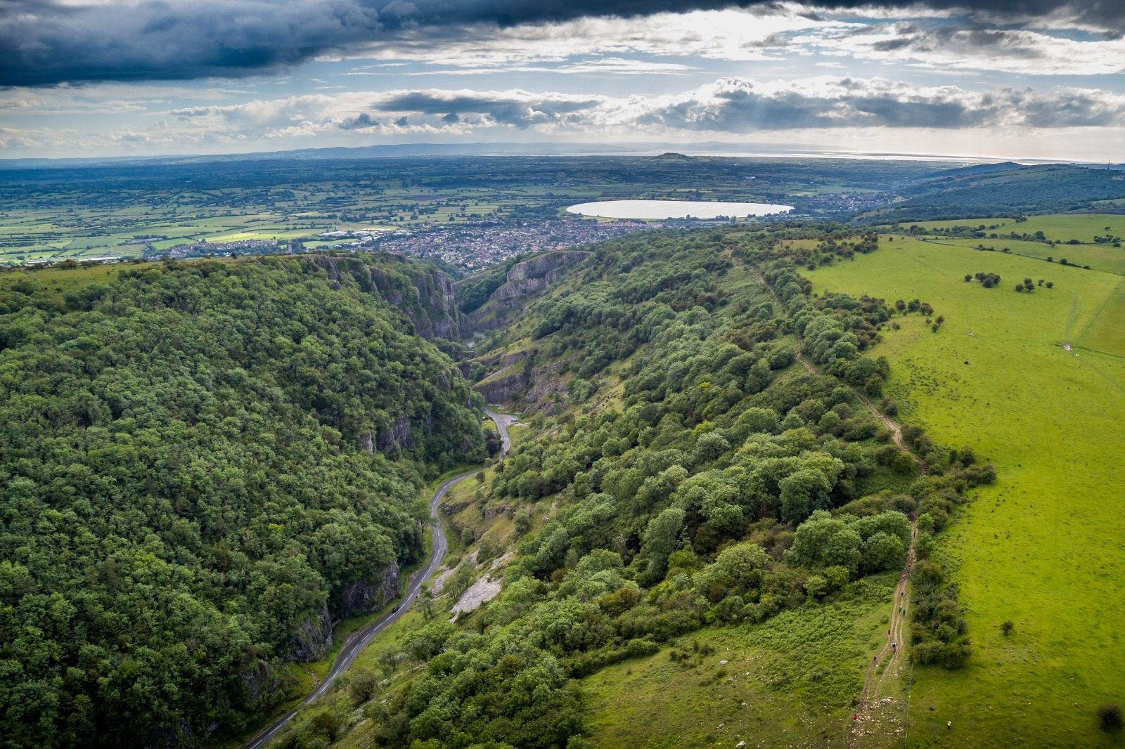 Aerial view of Cheddar Gorge, located in Somerset, England, featuring steep cliffs and lush greenery. A winding road is visible in the valley. Beyond the gorge, the landscape stretches into rolling hills and a distant town under a partly cloudy sky.