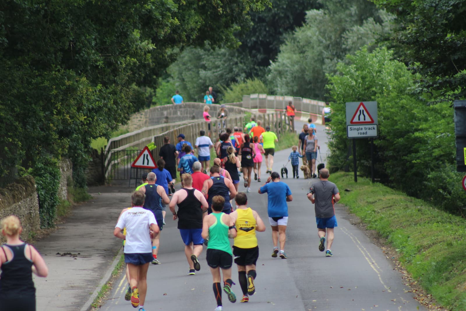 A group of people running on a paved road surrounded by greenery. Some runners are wearing bright clothing, and trees line both sides of the road. A sign on the right warns of a single track road ahead. A bridge is visible in the distance.