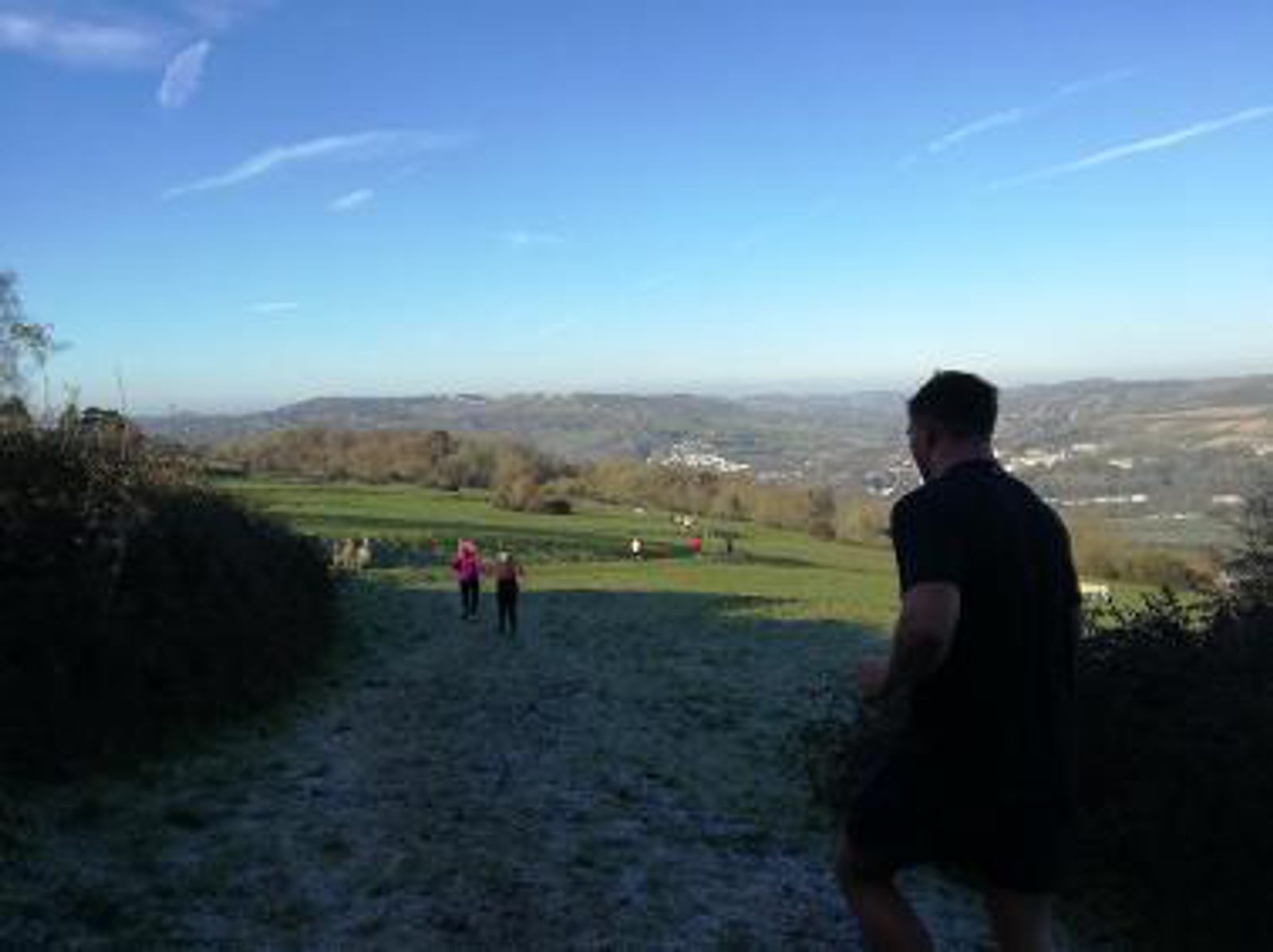 A person in a black shirt is running on a grassy hill with a scenic view of the countryside in the background. Trees and bushes line the path on the left, and several other people are seen walking ahead in the distance under a clear blue sky.