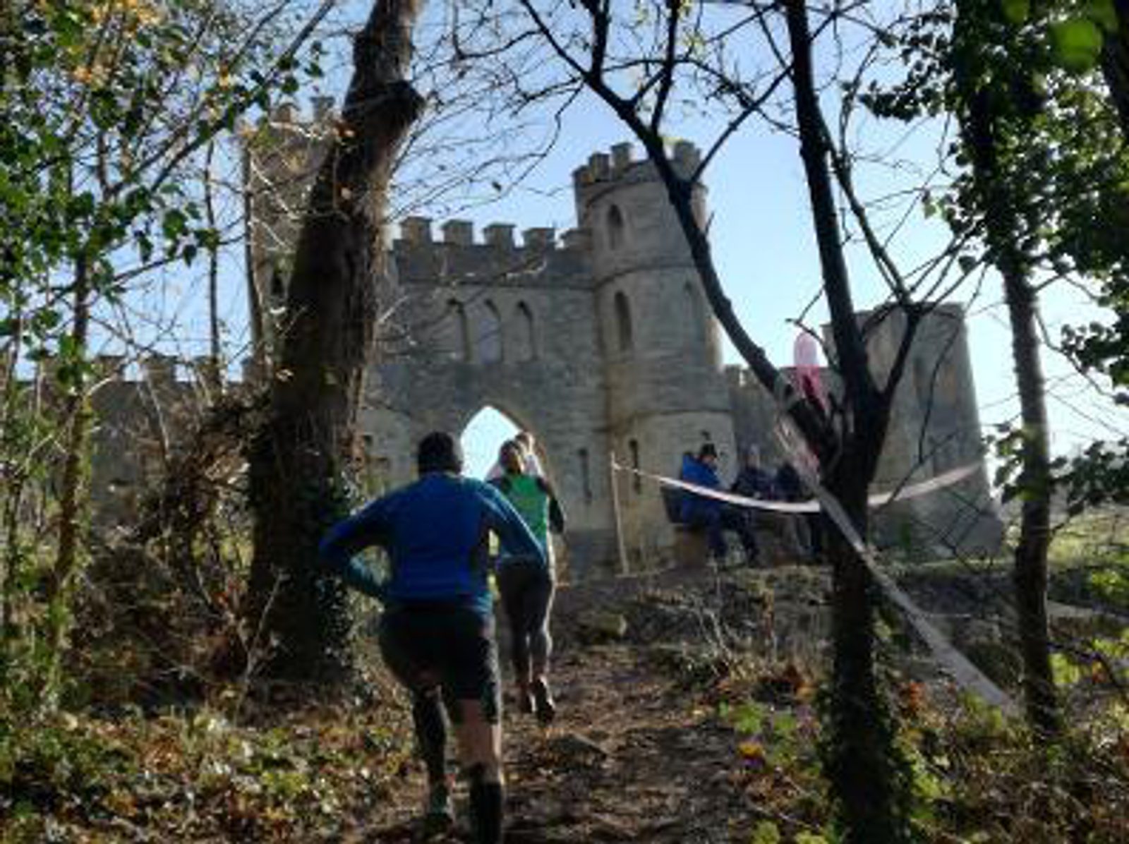Runners in sports gear jog towards a historic stone castle through a wooded trail, surrounded by trees and foliage. The castle features arched doorways and battlements, and a group of people can be seen standing near the entrance under a clear sky.