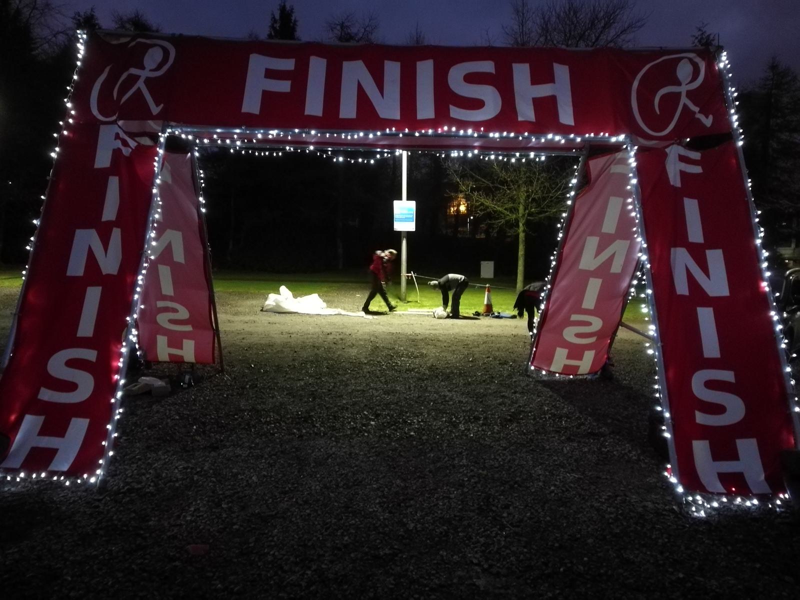 A red archway with the word "FINISH" written on it is illuminated with string lights in a dim outdoor setting. Several people are seen in the background near a signpost.